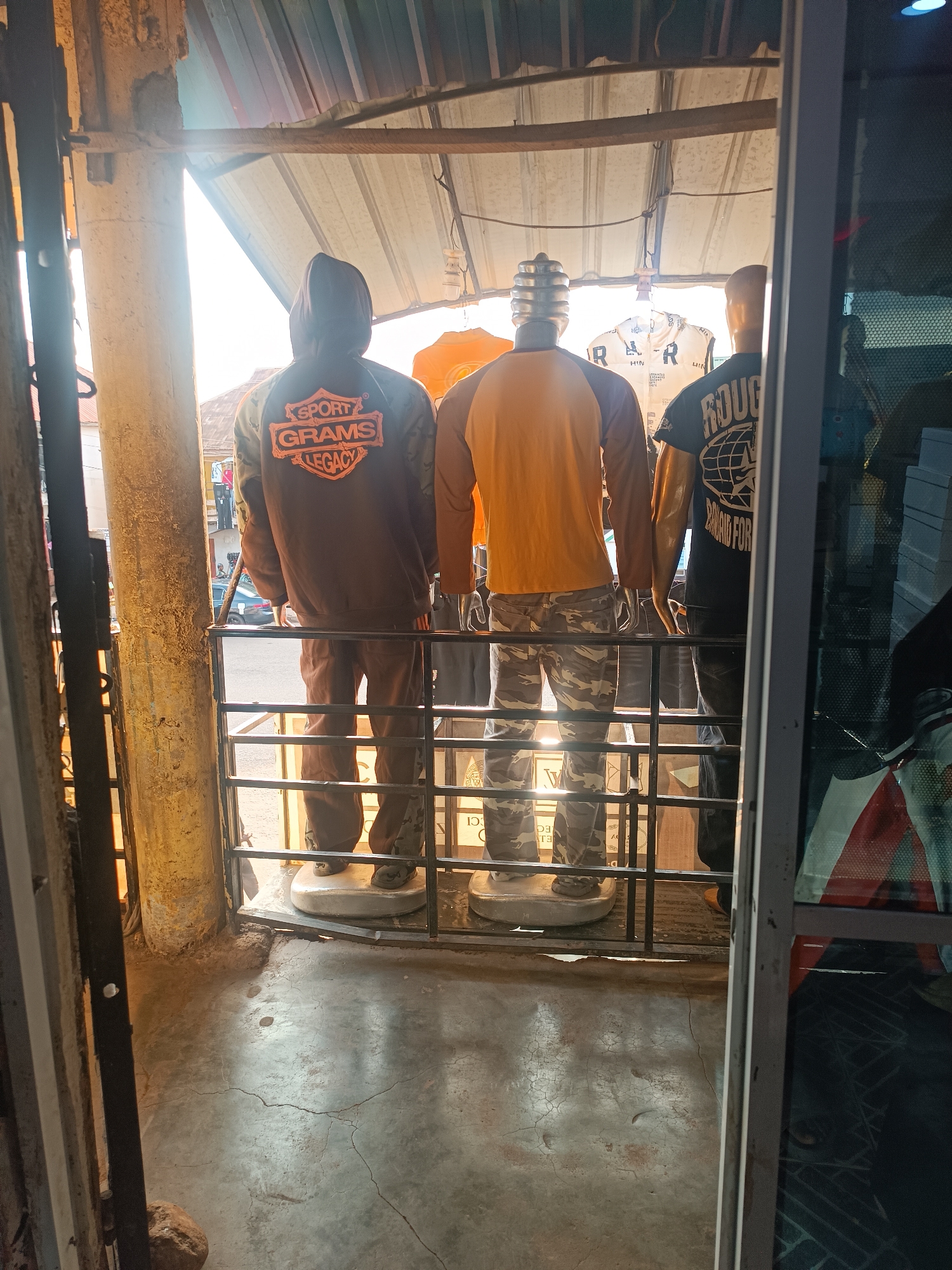 The image shows the back view of three mannequins standing behind a metal railing. The mannequin on the left wears a brown hooded jacket with "SPORT GRAMS LEGACY" printed in orange. It also wears matching brown pants. The middle mannequin wears a mustard-yellow long-sleeved shirt and camouflage pants. The mannequin on the right wears a black t-shirt with "ROUGH DIAMOND FOR..." printed on it and black pants. The mannequins stand on round white bases. The scene is in a storefront, viewed from the outside. The floor is concrete and cracked. The location is Bukuru, Nigeria.