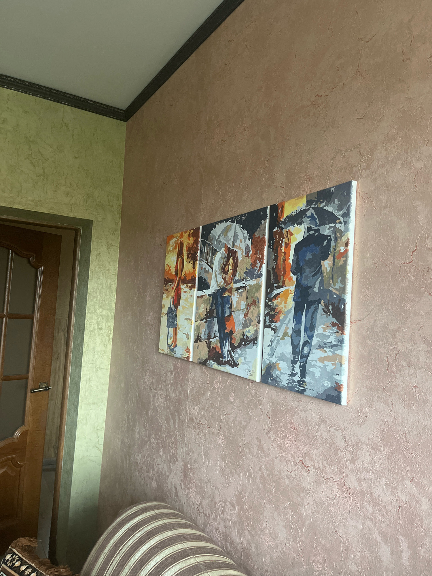 A three-panel abstract painting of a city street scene in the rain is mounted on a textured, light brown wall. The painting depicts figures holding umbrellas and walking through wet, reflective streets. To the left of the painting is a doorway with a wooden door with glass panes. The door is partially open, revealing a hallway or adjacent room with a light green textured wall. The ceiling above is white with dark molding along the edge. In the foreground, the striped upholstery of a sofa is visible. The overall environment appears to be an indoor residential space, likely a living room or hallway, in Vladimirovka, Russia.