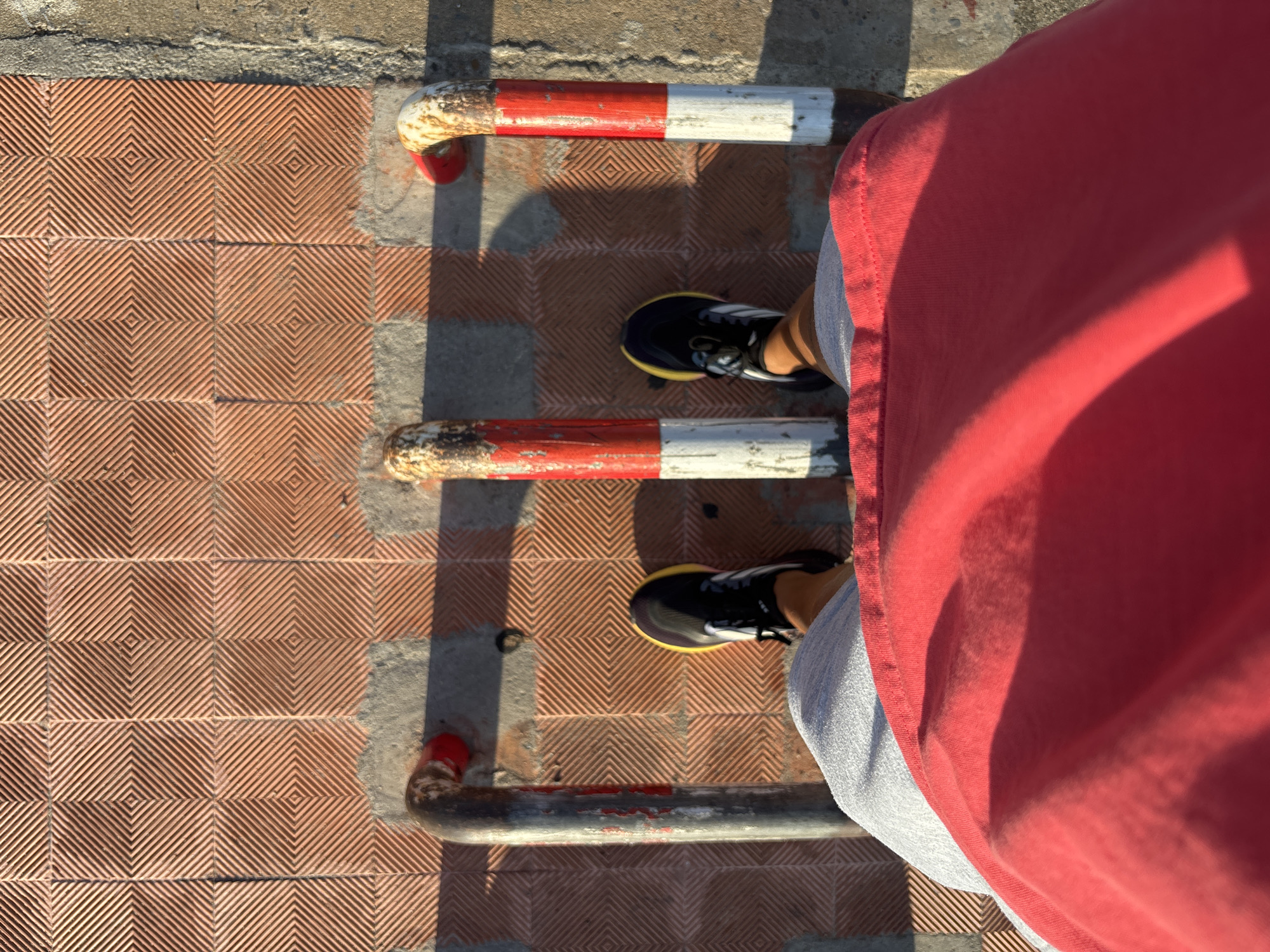 A first-person perspective shot from a person wearing gray shorts and a red t-shirt, standing on a patterned red tile walkway. In front of the person are three metal posts, painted with alternating red and white stripes. The ground is a mix of the red tiles and a rough gray concrete surface. The lighting suggests it is daytime, with shadows cast by the posts and the person's legs. The location is identified as Bac Nha Trang Ward, Vietnam.
