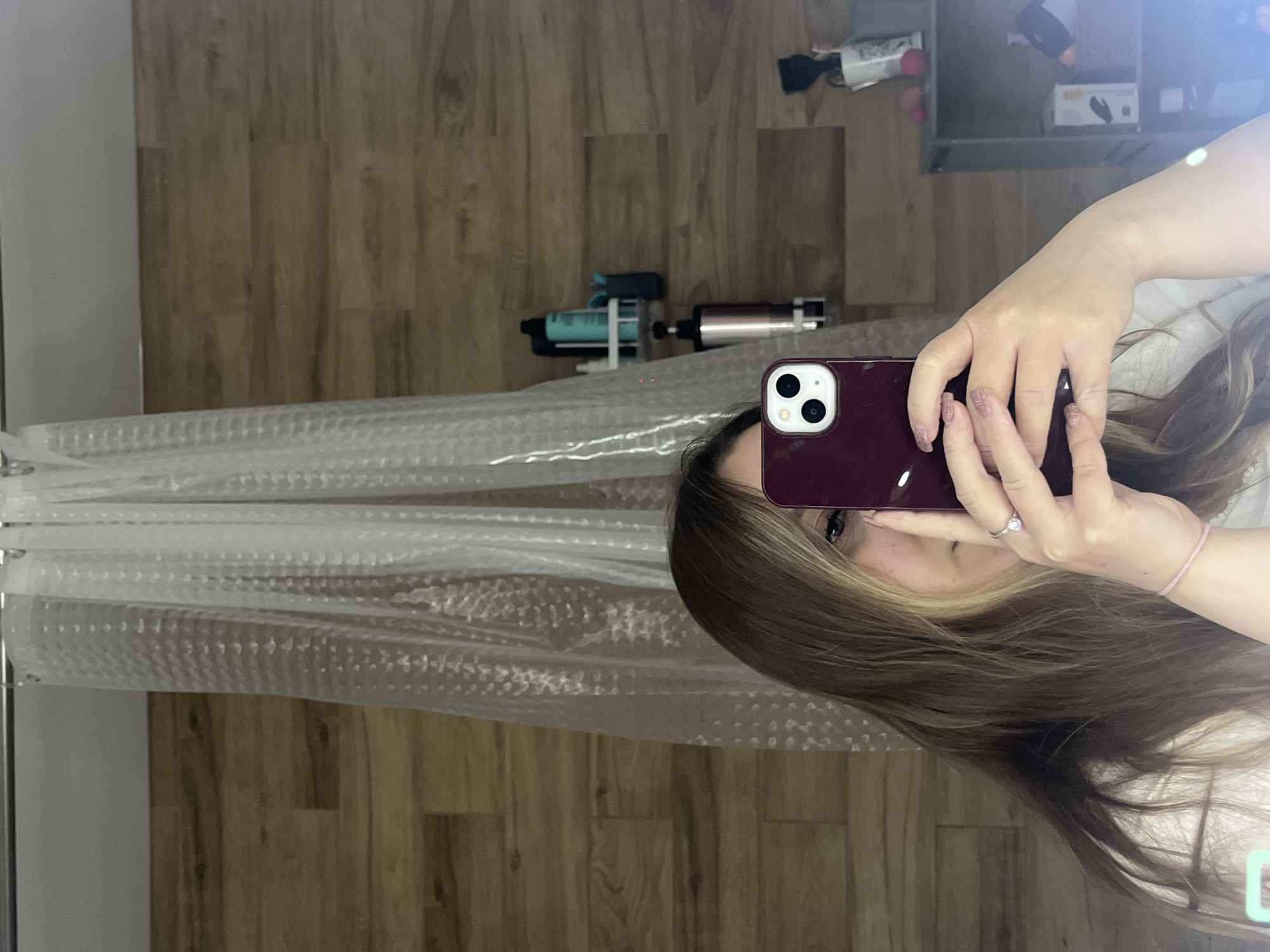 A person takes a selfie in a bathroom. The bathroom has a shower curtain, wood-look flooring, and a shelf with various items. The person holds a burgundy smartphone. The location in Krasnodar, Russia, cannot be determined from the image.