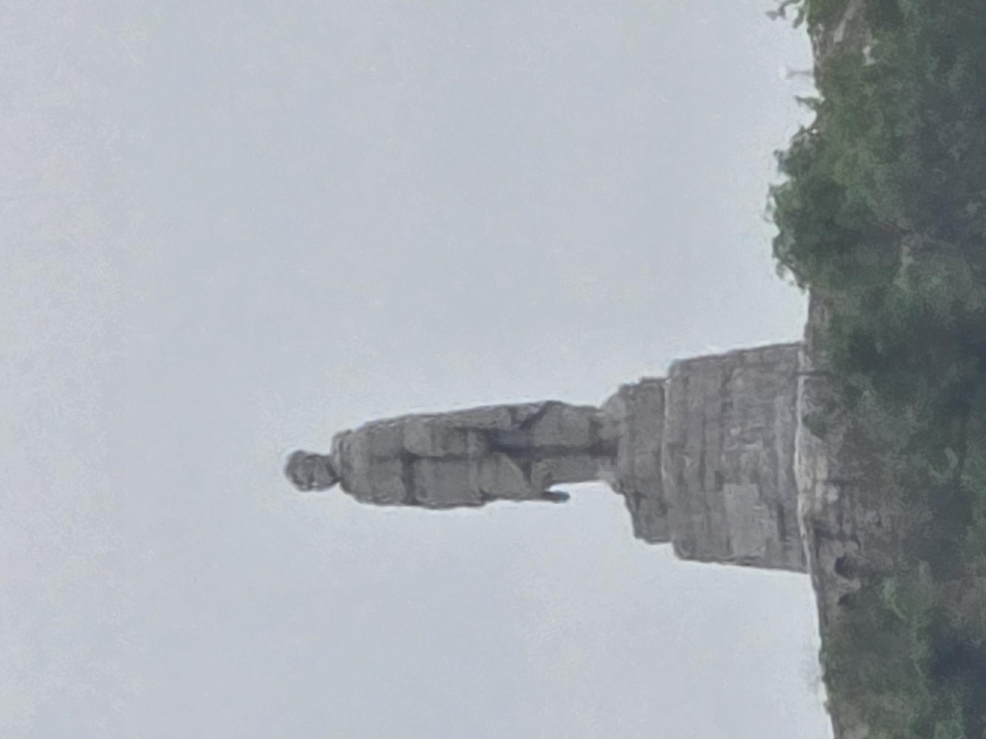 A horizontal photograph depicts a rock formation on a cliff face. The formation resembles a person seated, and it protrudes from the main cliff edge. Some greenery grows on the cliff face to the right. The background consists of an overcast sky. The location is the Hang Tom viewpoint in Mu Cang Chai, Vietnam.