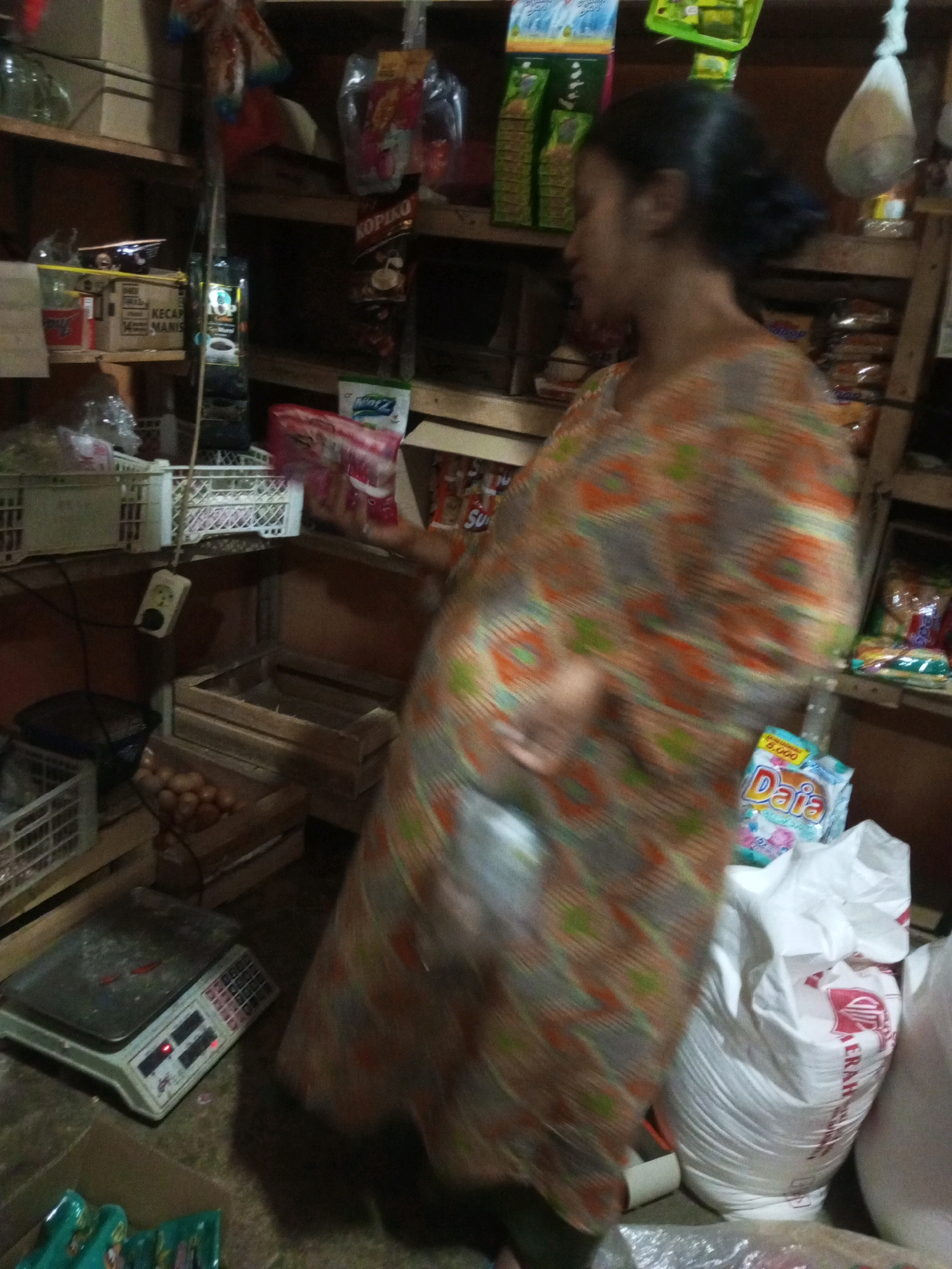 The image depicts an indoor scene, likely a small shop or market stall in Andongsili, Indonesia. A woman, facing away from the camera, is visible in the foreground, wearing a patterned orange and green garment. She appears to be reaching towards or holding a white plastic bag.
The environment is cluttered with various goods displayed on wooden shelves and crates. On the shelves, there are packaged food items, including what appears to be coffee (Kopiko) and other snacks. Below the shelves, a digital weighing scale with a red indicator is prominent on the floor. Several wooden crates are stacked, one of which contains a pile of brown eggs. Large white plastic bags, some with red lettering (likely indicating a brand name like "MERAH"), are situated on the floor near the woman. The lighting in the scene is dim, suggesting an indoor, possibly low-light environment. The overall impression is of a busy, utilitarian retail space.