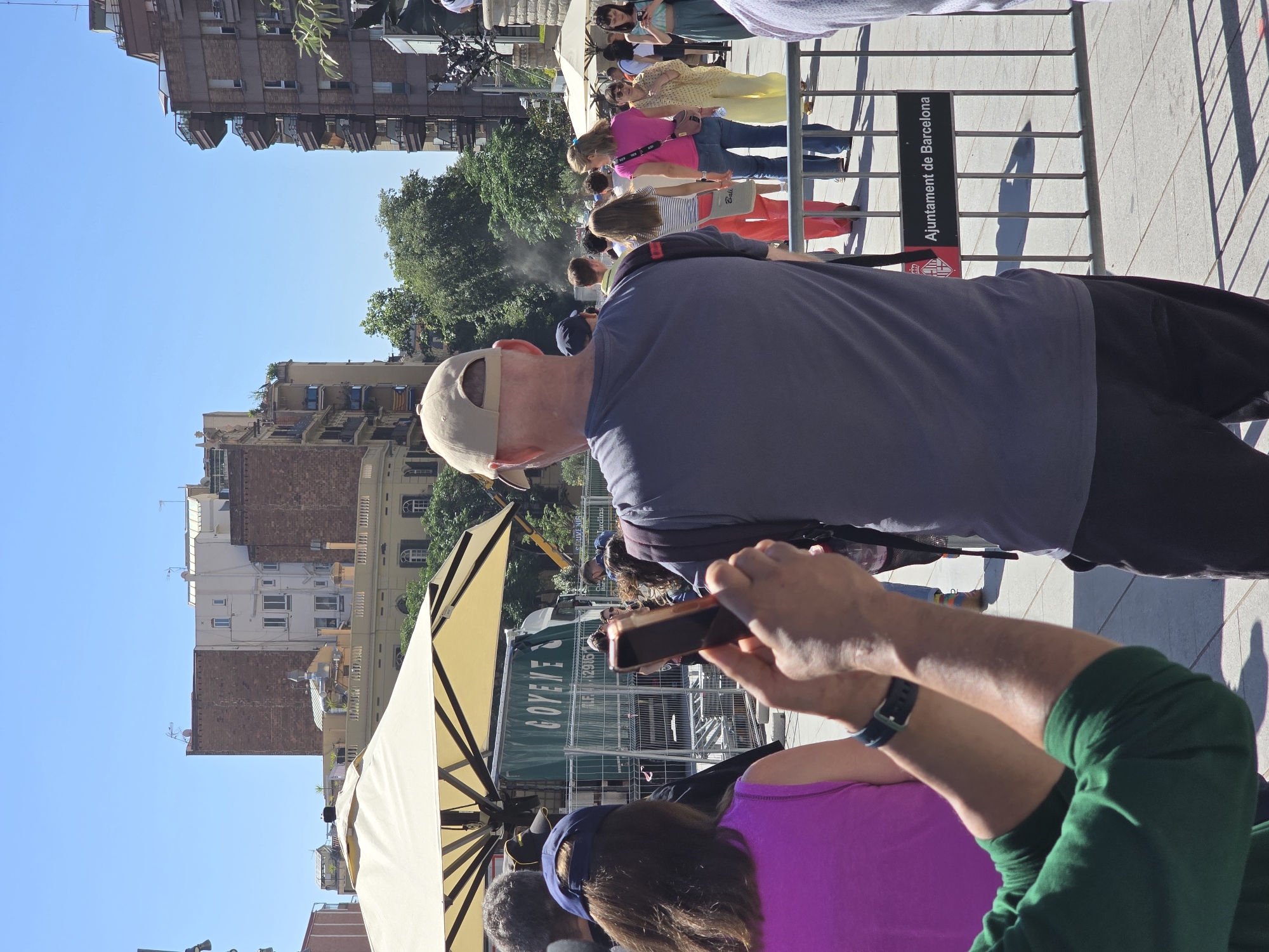 This is a medium-distance, eye-level, outdoor, daylight, eye-level shot of a crowd of people in Barcelona, Spain. The image is taken from behind some individuals, looking towards a street scene with buildings and trees in the background. In the foreground, a person wearing a green long-sleeved shirt and a blue wristband is holding up a mobile phone to take a photo. To the right and slightly further back, a person in a grey t-shirt and black pants is also facing away from the camera, appearing to be watching something in the distance. A woman in a bright pink top and blue jeans is visible further back, as is another woman in a yellow outfit and a woman in a blue skirt. Behind them, a fence with a sign reading "Ajuntament de Barcelona" is visible. A green truck with the word "GOYENE" on its side is parked behind a temporary fence, covered by a large tan umbrella. The background shows typical Barcelona architecture with brick and stucco buildings under a clear blue sky. The scene appears to be a public space, possibly a street or plaza, with people engaged in various activities like taking pictures and observing.