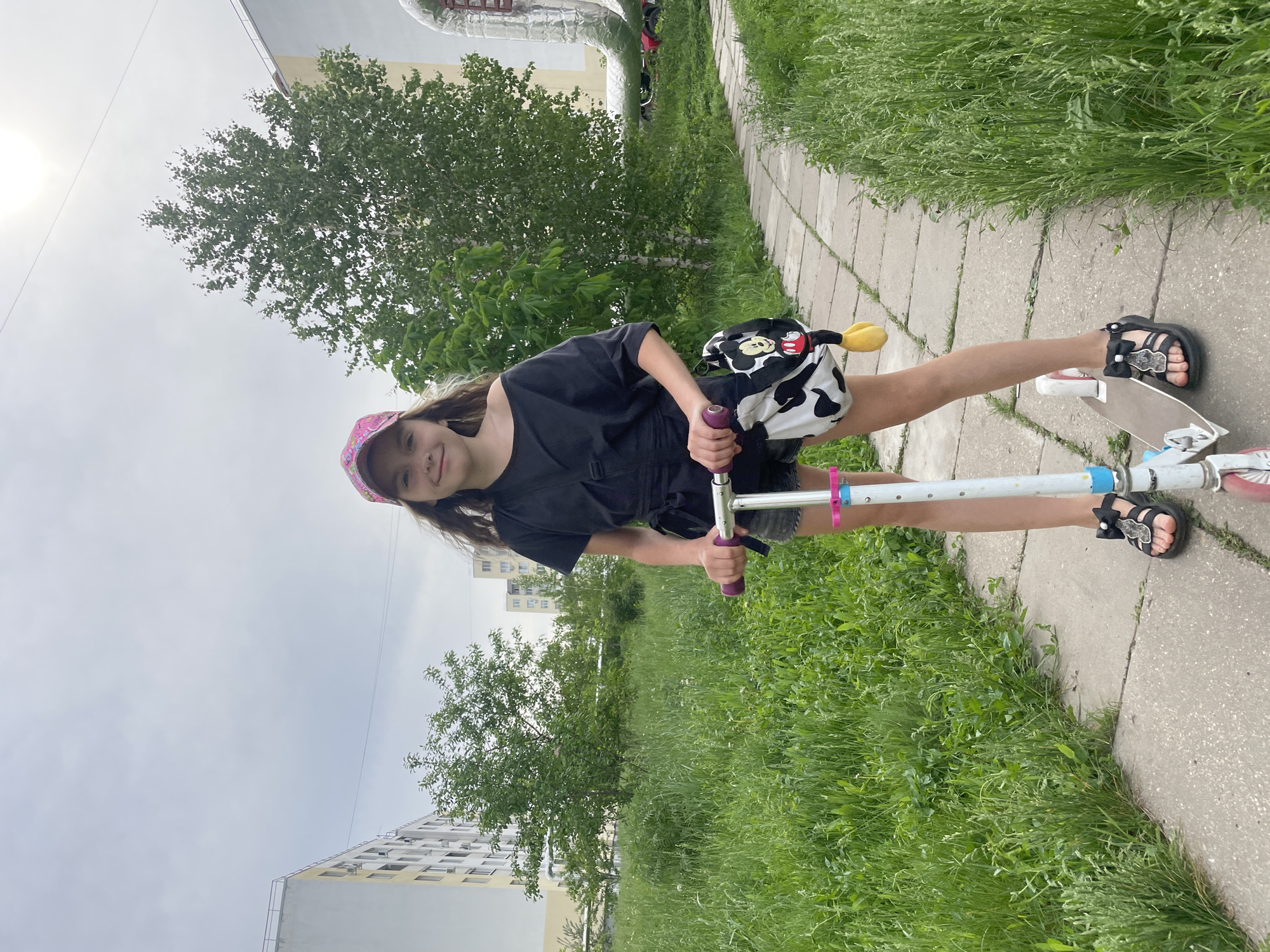 A young girl stands on a scooter on a paved path in what appears to be a park-like area within Saratov, Russia. She is wearing a black t-shirt, dark shorts, and black sandals. A pink cap is on her head, and she has a backpack with a Mickey Mouse toy attached. The scooter is white and appears to be worn, with purple grips. The girl is smiling at the camera. To the left and right of the path, there is green grass. In the background, buildings and trees are visible. The sky is overcast.