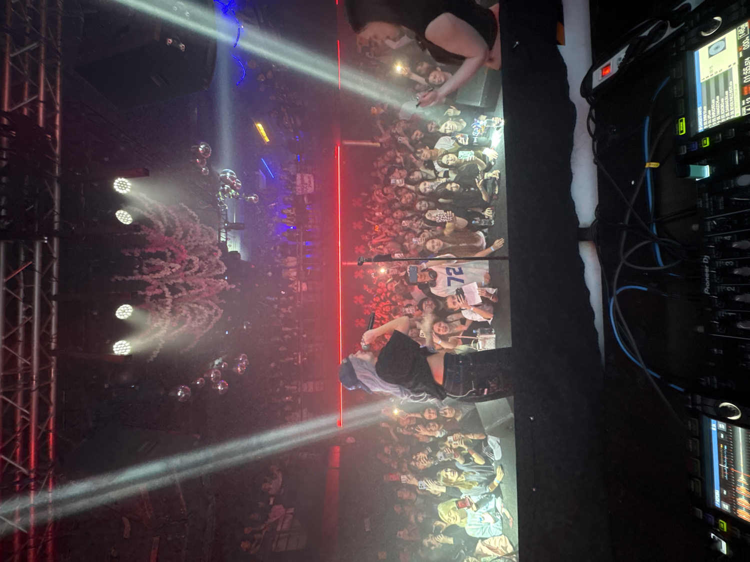 This image captures a vibrant, high-energy indoor concert or performance. A performer, with light-colored hair and dressed in dark attire, is visible towards the bottom center of the frame (when viewed with correct orientation), holding a microphone and interacting with the audience. A large crowd of people fills the background, looking towards the stage. Many audience members are holding up smartphones, likely recording the event, and their faces convey excitement and engagement. One person in the crowd wears a shirt displaying the number "72."
The stage area is illuminated by multiple bright spotlights that pierce through visible haze or smoke, creating distinct light beams. Several mirrored disco balls hang, reflecting light and contributing to the dynamic atmosphere. A prominent red linear light strip runs horizontally across the stage's backdrop. On the left side, a burst of pink or red confetti or similar particles is captured mid-air, suggesting a special effect during the performance. Metal truss structures, supporting lighting and possibly speakers, are also visible on the left.
To the far right, a DJ booth or sound engineering station is present, equipped with Pioneer DJ mixers and media players. Numerous blue and black cables connect the equipment. Screens on the DJ gear display waveforms and numerical information, including "01.55.94" and "41.13." The overall setting is dark, characteristic of a nighttime event in a performance venue in Vladivostok, Russia, highlighting the stage lighting and audience interaction. The original image is rotated 90 degrees clockwise from its natural upright orientation.