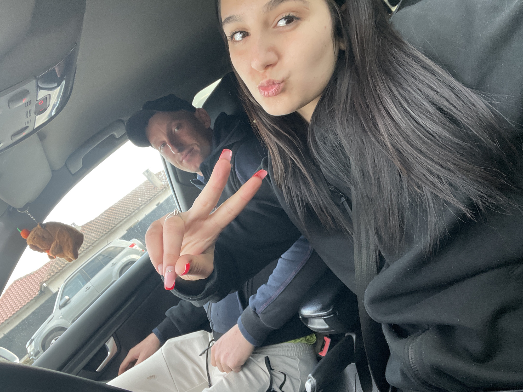The image appears to be taken inside a car. A young woman with long, dark hair and red nail polish is in the foreground, making a kissing face and holding up the peace sign. Behind her is a man with short hair, wearing a black baseball cap and looking towards the camera. The man is wearing white pants. Outside the car, a residential area is visible with houses and another car parked on the street. A plush brown animal with an attached orange carrot is hanging from the rearview mirror. The location is indicated by the prompt to be Kavarna, Bulgaria.