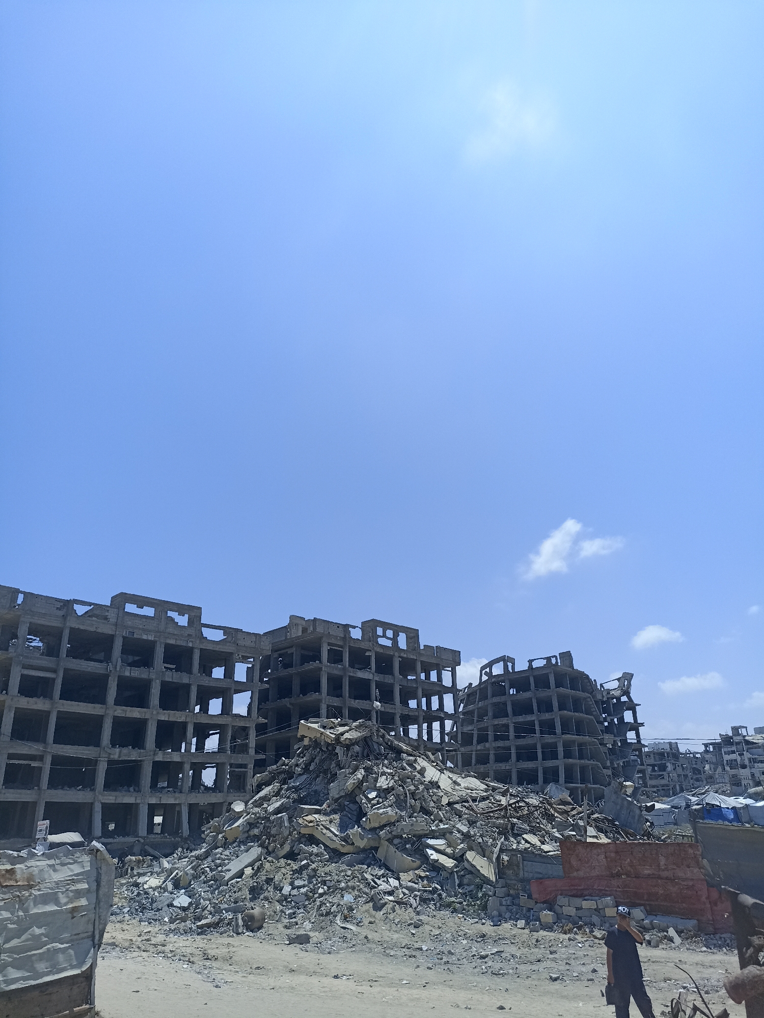 The image depicts a scene of devastation in Jabalia, Palestinian Territories, under a clear blue sky with scattered clouds. In the foreground, a large pile of rubble dominates the center of the frame, consisting of concrete debris, twisted metal, and fragmented building materials. To the left and behind the rubble, the skeletal remains of several multi-story concrete buildings are visible, their floors and walls exposed and damaged, indicating extensive destruction. Some of these structures appear to be partially constructed or heavily damaged, with rows of empty window frames and exposed rebar.
In the far right of the image, a person wearing a black t-shirt and dark pants stands facing left, with their right hand near their face, possibly adjusting a cap or mask. They are standing on a dusty, uneven ground covered with smaller debris. A red tarp or barrier is visible behind the person, and further in the background, more damaged structures and some makeshift tents or shelters can be seen. The overall environment is one of widespread destruction and aftermath.