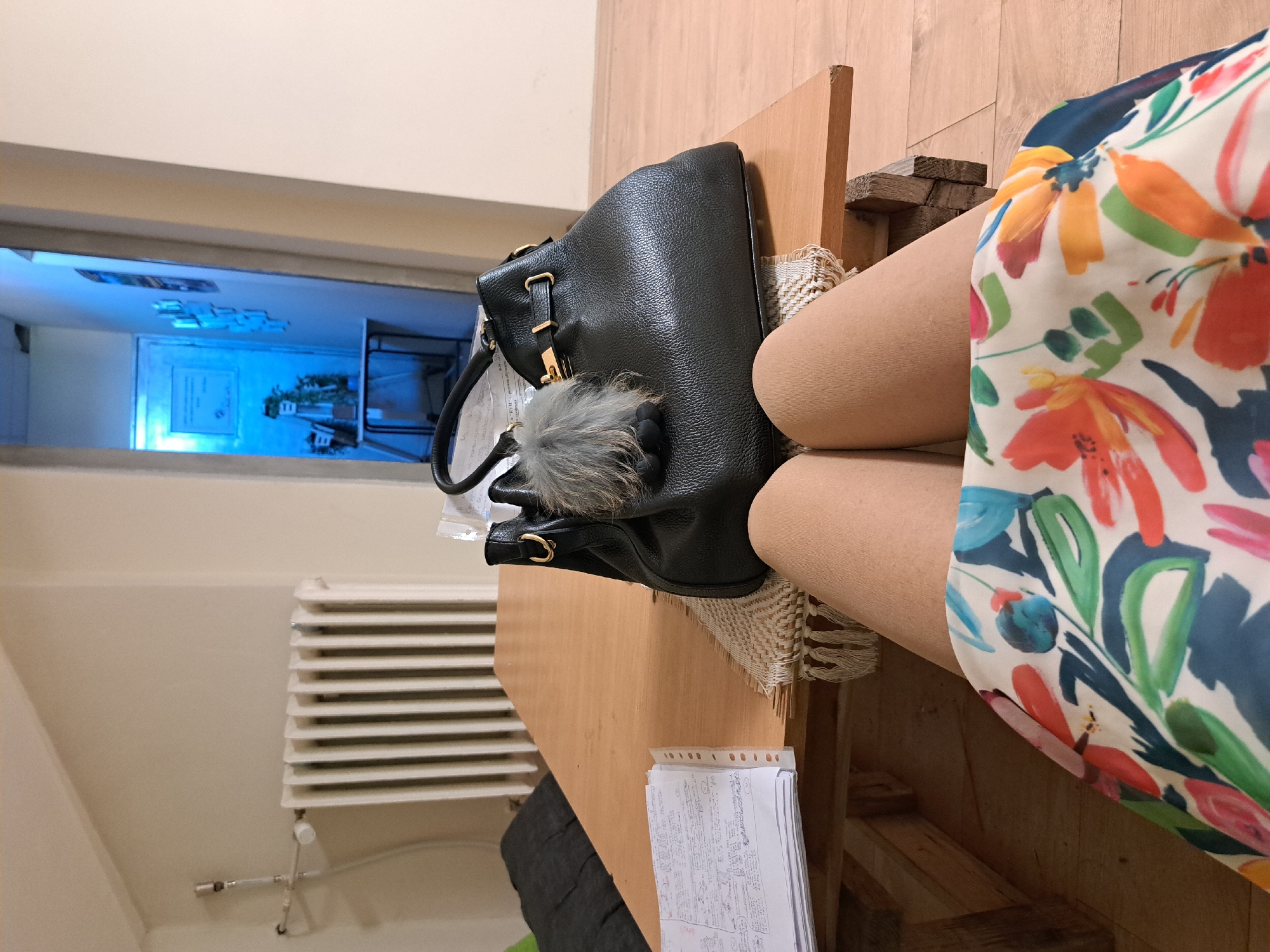 The image depicts a scene indoors, likely a room in a house or apartment. A person wearing a floral print dress sits at a wooden table. A black leather handbag with a grey fur pom-pom rests on the table next to them. A stack of papers is also on the table. In the background, a radiator can be seen on the wall. Through a window, the interior of a building is visible. The specific location within Sofia, Bulgaria cannot be determined from the image.