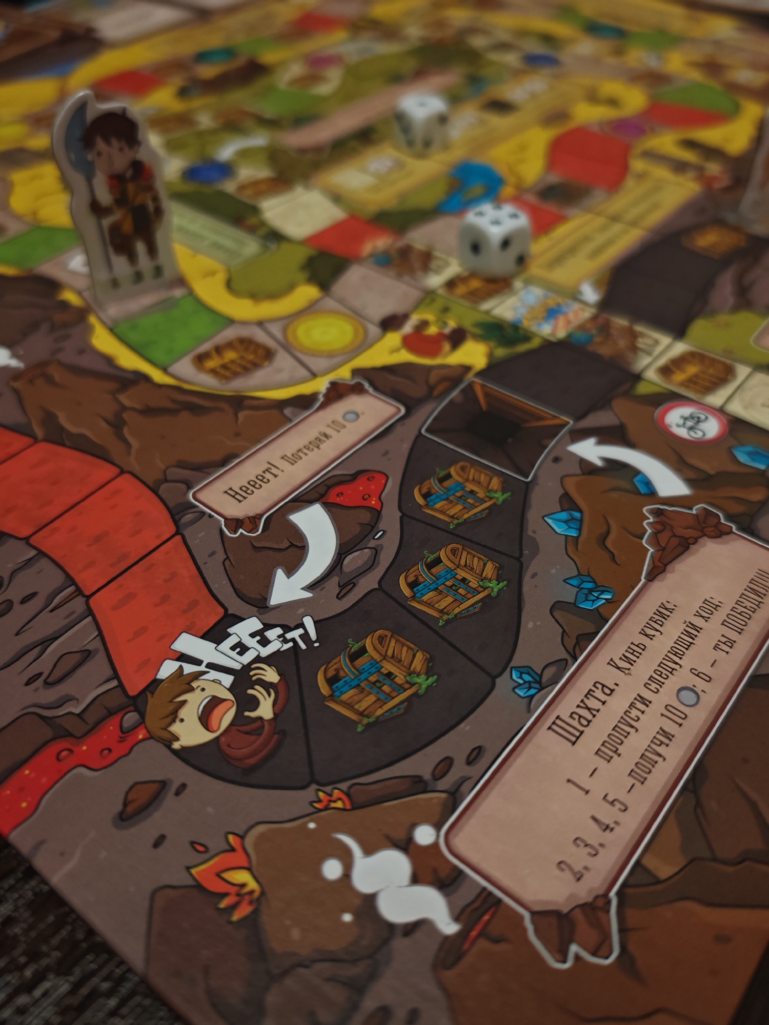 The image shows a close-up view of a board game with a mining theme. The board is laid out with a winding path, represented by red bricks, leading through a rocky terrain with brown and yellow elements. Several game pieces are visible, including a small character figure standing on a yellow space. There are also dice on the board, one white with black dots showing a 1 and another white with black dots showing a 6. Two separate rectangular cards are placed on the board. One card has text that appears to be in Russian, reading "Нееет! Потеряй 10." (Neeet! Lose 10.) and features a red splat with white dots. The other card, also with Russian text, reads "Шахта. Кинь кубик: 1 — пропусти следующий ход; 2, 3, 4, 5 — получи 10; 6 — ты победишь!" (Shahta. Roll the die: 1 — skip next turn; 2, 3, 4, 5 — get 10; 6 — you win!). This card is positioned near several illustrated treasure chests. The overall scene is a detailed shot of a board game in progress, with elements suggesting movement, chance, and objectives within the game. The lighting is natural, and the focus is sharp on the foreground elements, with the background becoming progressively blurred.
