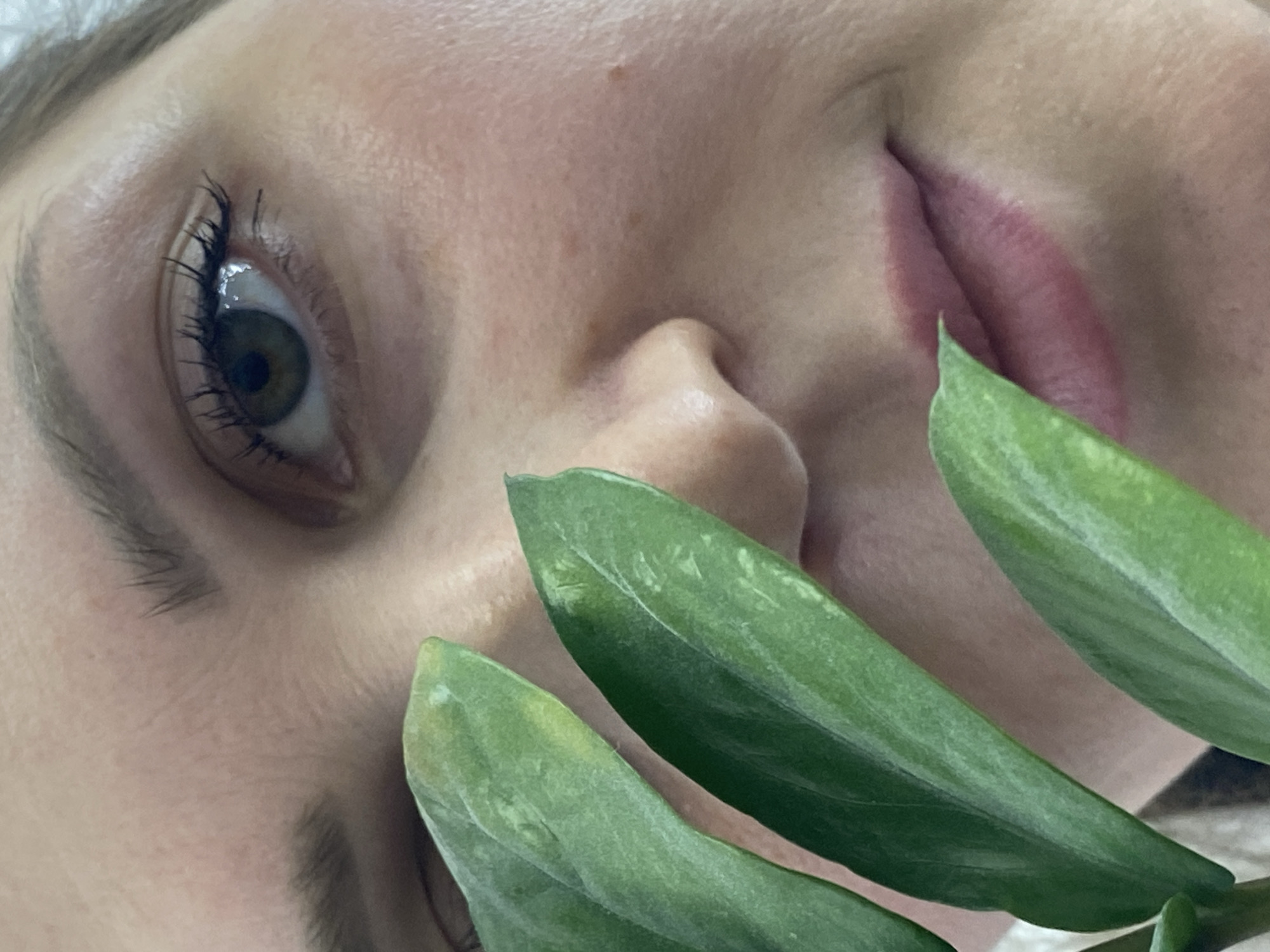 A close-up, tilted image features a fair-skinned person with their eye visible. The person has light brown eyebrows and dark eyelashes. Their nose and mouth are partially visible, with pink lips. Three green leaves are positioned in front of the person's face, partially obscuring it. The background is out of focus. The location cannot be confirmed to be in Марковский, Russia based on the visual information.