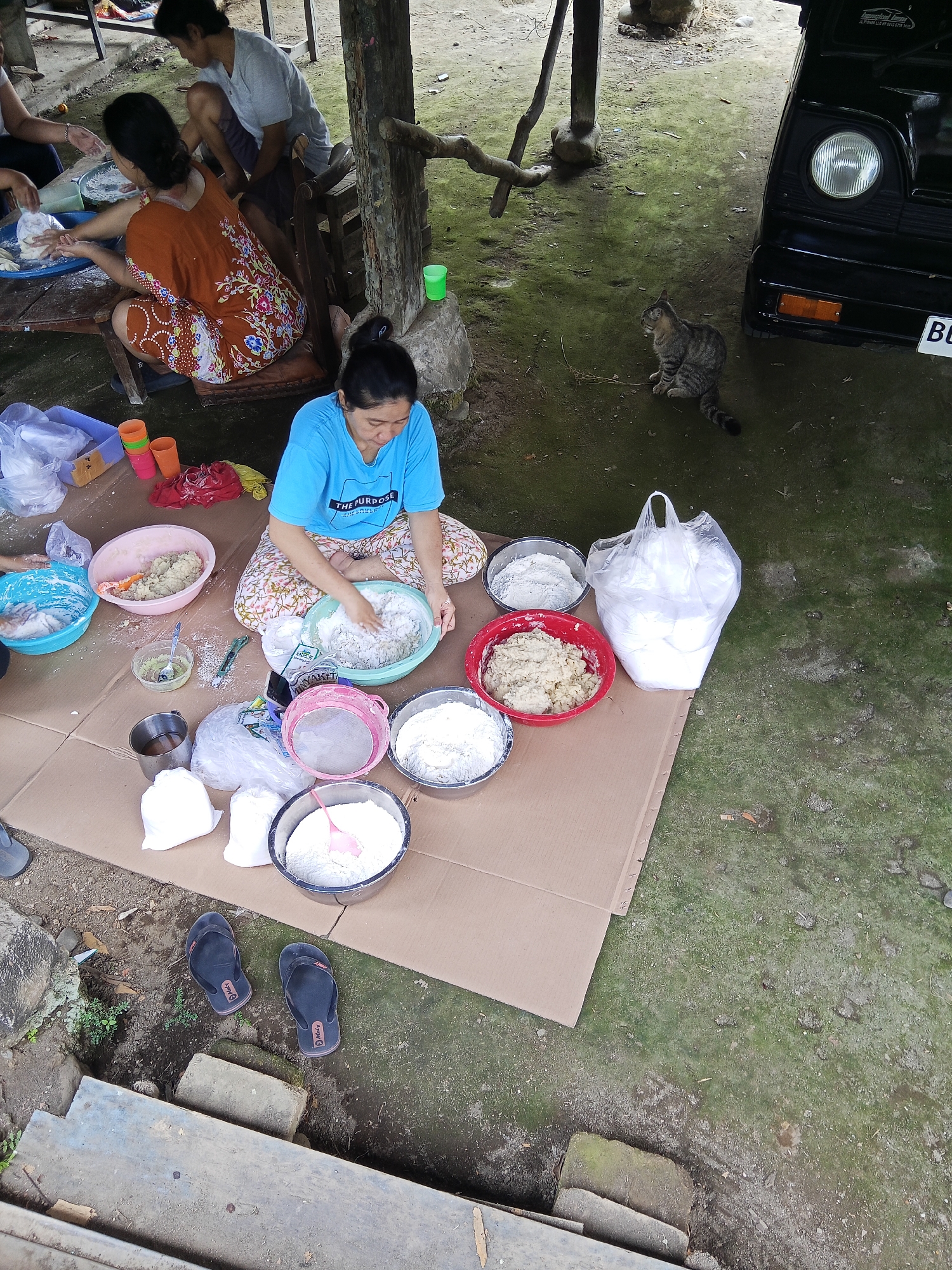 In an outdoor setting, likely in Lubuklinggau, Indonesia, several individuals are engaged in food preparation. The ground is covered with a large piece of cardboard, upon which numerous bowls and containers are arranged. One woman, wearing a blue t-shirt with text, is actively mixing ingredients in a green bowl with her hands. Another woman, dressed in an orange patterned top, is also working with food in a blue plastic basin. A third person, partially visible, is wearing a grey t-shirt.
Scattered around the preparation area are various ingredients and tools, including bowls of flour, what appears to be dough, a sieve, and bags of white powder. Several plastic cups and a metal container are also present. In the background, a black vehicle is parked, and a tabby cat is visible on the ground nearby. The environment appears to be somewhat rustic, with exposed wooden beams and green, mossy ground, suggesting an informal, possibly rural, setting within the city. Two pairs of flip-flops are discarded on the ground near the edge of the cardboard.