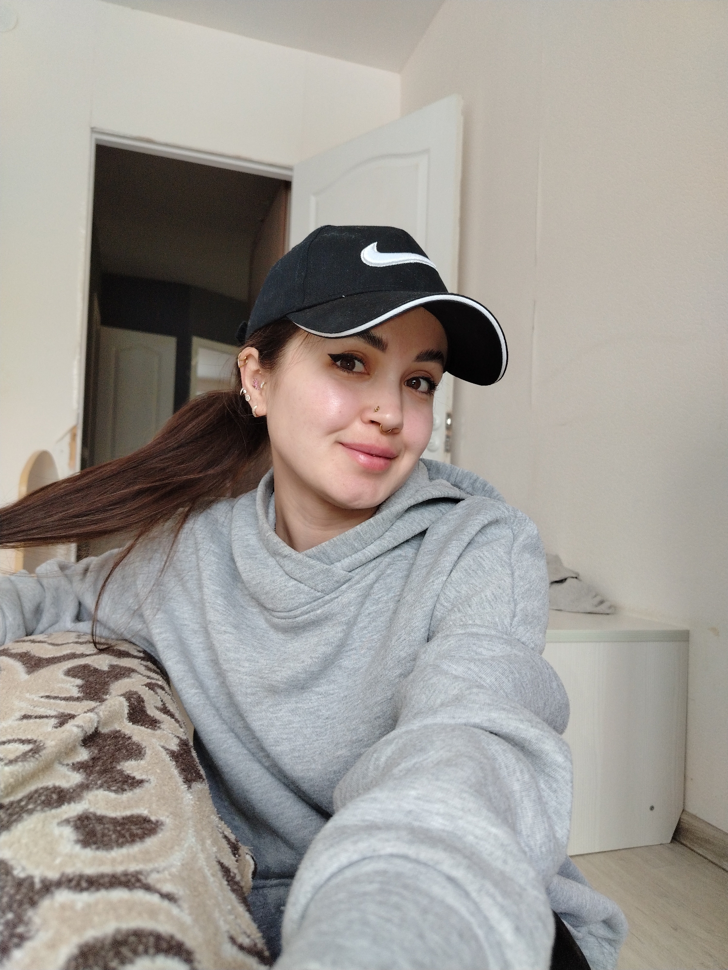 A medium shot shows a young woman with fair skin, wearing a black baseball cap with a white Nike logo, posing for a selfie in an indoor setting. Her dark hair is pulled back into a ponytail. She is wearing a gray hooded sweatshirt. She has multiple piercings in her left ear and a nose ring. The background includes a doorway with a partially opened white door and a white cabinet. The city and state are not discernible.
