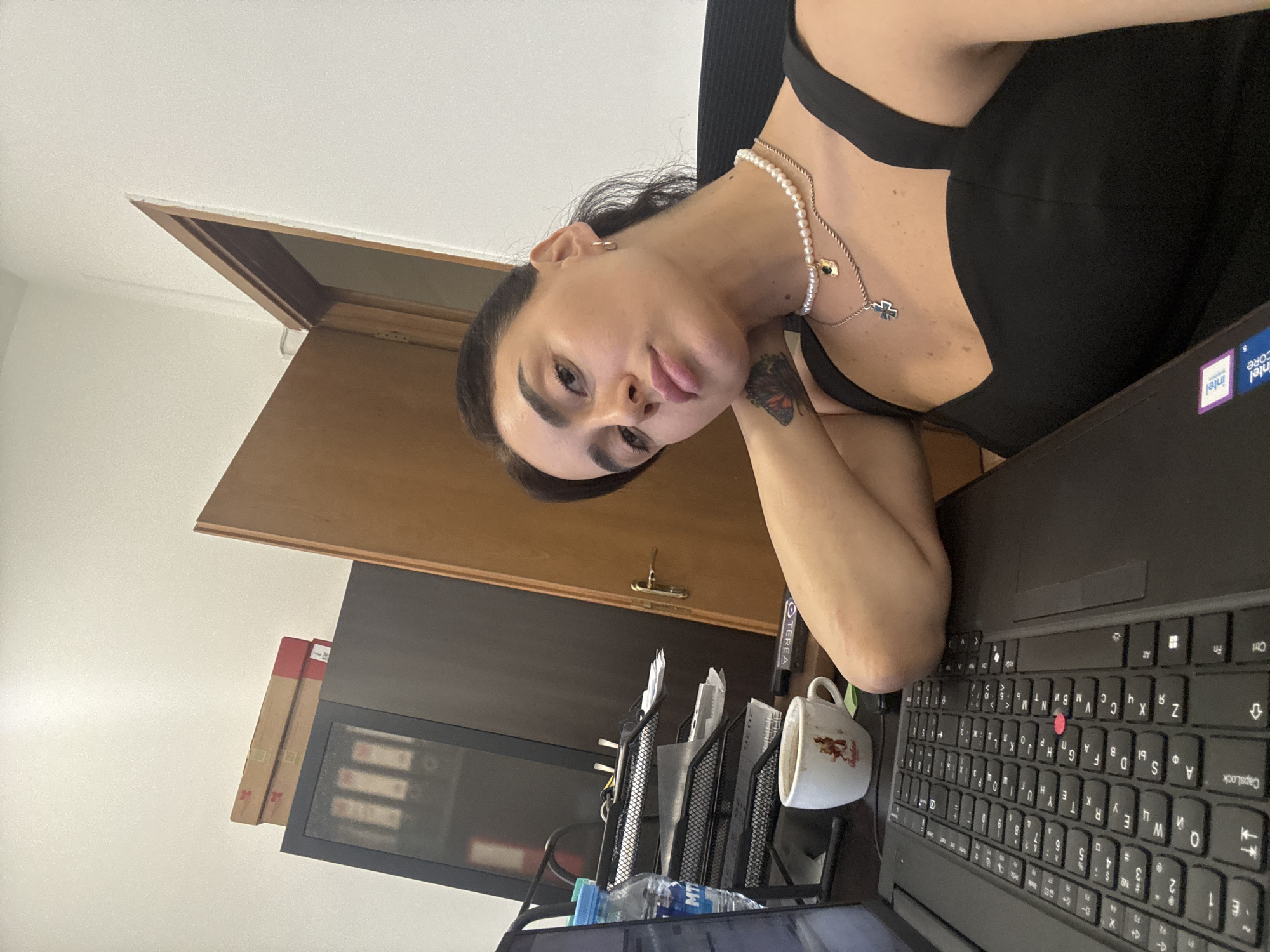 The image depicts a young woman sitting at a desk in what appears to be an office environment in Tbilisi, Georgia. She is positioned in the foreground, leaning her head on her left hand, with her right arm resting on a laptop. The woman has dark hair pulled back and is wearing a black dress and pearl necklaces.
The desk is cluttered with office supplies, including a laptop, a cup of coffee, a water bottle, and a document organizer. In the background, a wooden door with a golden handle is visible, as is a dark-colored cabinet filled with binders and boxes. The lighting suggests an indoor setting, likely during daylight hours. The overall scene conveys a sense of work or study in a typical office space.