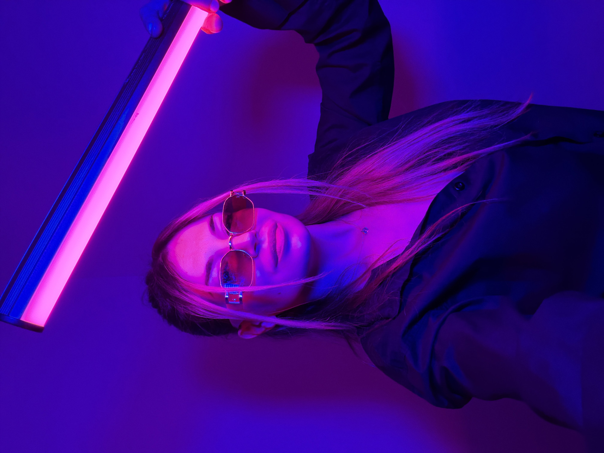 A stylized, high-contrast portrait features a person, likely female, positioned horizontally across the frame. The subject wears dark, reflective sunglasses and a dark garment, possibly a jacket or shirt. Her light-colored hair, appearing vibrant pink under the dramatic lighting, flows around her head and shoulder.
A prominent glowing light tube, emitting an intense neon pink light, is held by the subject's hand in the upper left of the image. This light source directly illuminates her face, neck, and hair, casting strong pink hues and creating defined shadows. The background is a solid, deep blue-purple color, providing a stark, moody contrast to the bright pink light. The dark clothing takes on blue and purple tones from the ambient lighting.
The overall aesthetic is futuristic and enigmatic, dominated by electric pink, various shades of purple, and deep blue. The subject's expression is neutral, obscured slightly by the sunglasses. The composition is dynamic, with the diagonal light tube cutting across the frame and the subject's horizontal pose. The artificial lighting suggests an indoor studio setting or a nighttime shoot. No text is visible in the image. The scene is set in Budva, Montenegro.
