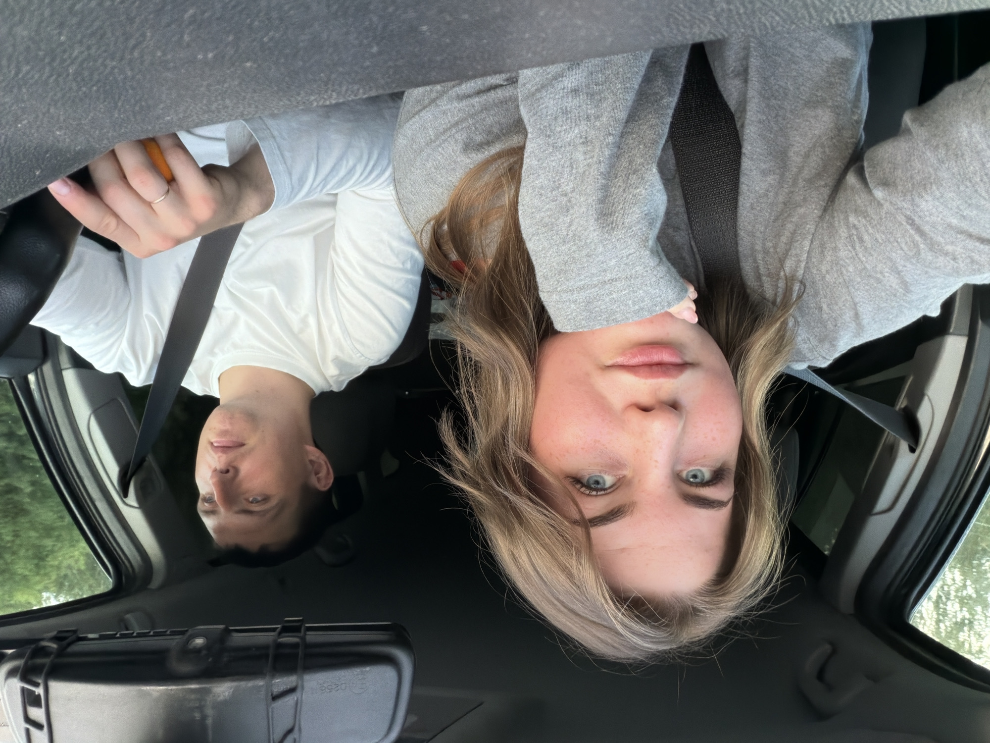 Two individuals are visible inside a vehicle. The woman has blonde hair, blue eyes, fair skin, and is wearing a grey jacket. The man has short dark hair and fair skin, and is wearing a white shirt. Both individuals are wearing seatbelts. The man is holding a round, orange object in his hand. A black rectangular case is in the lower portion of the frame. The setting appears to be the interior of a car, possibly on a road trip. The scene is not explicitly located in Belaya Gora, Russia; no geographic features or signs indicate that specific location.