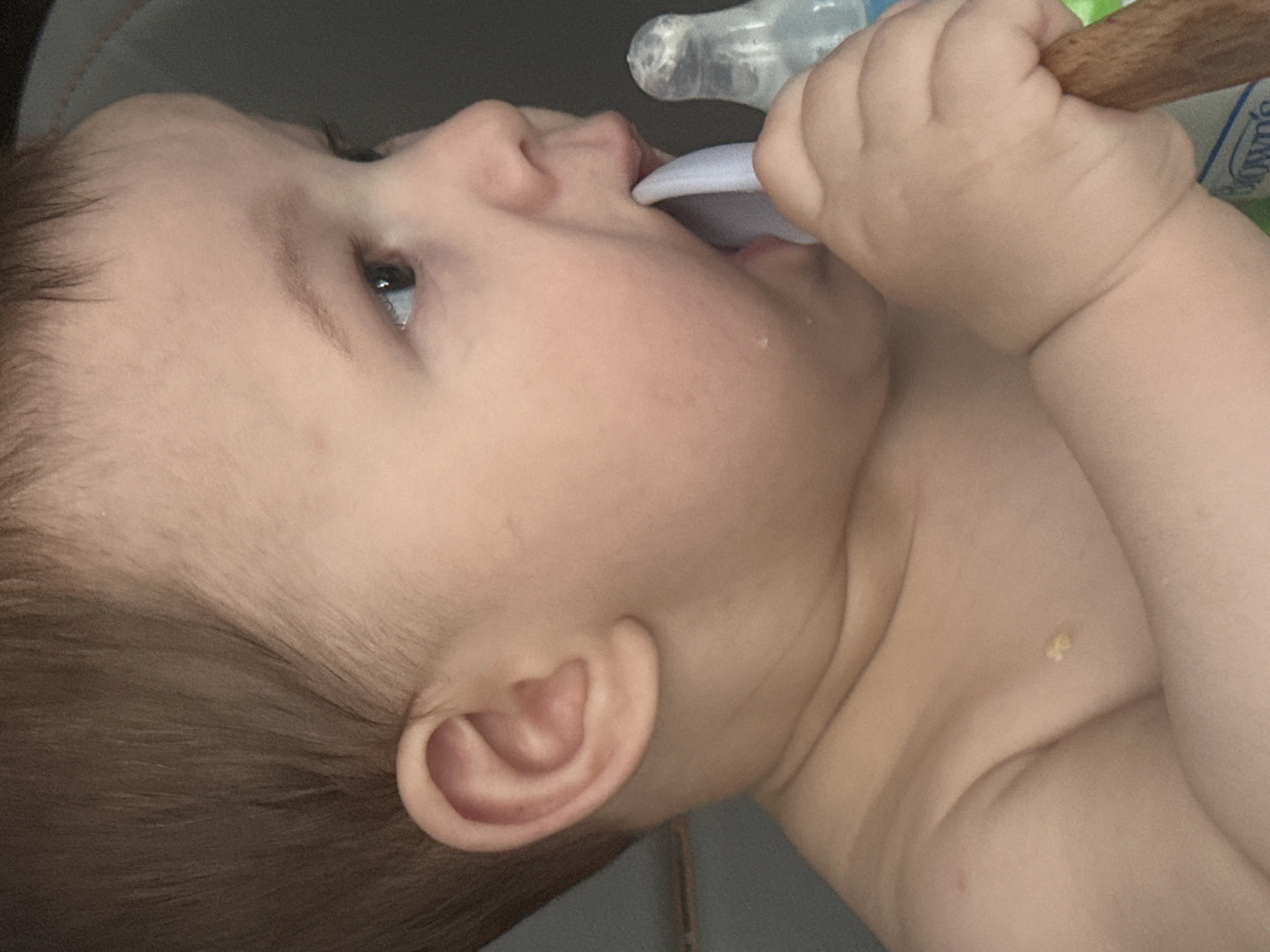 A close-up, profile shot shows a baby, viewed from the right side, holding a wooden spoon in their mouth and a plastic bottle to their right. The baby's left ear is visible on the left side of the frame, and their right eye is looking to the right. The baby's skin is fair, and they have brown hair. The background is a neutral gray.