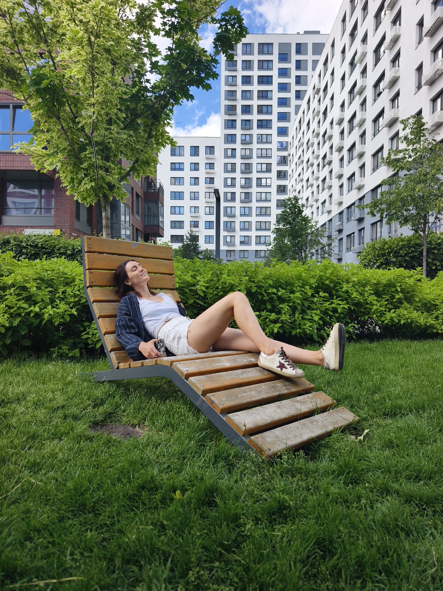 A young woman is resting on a wooden lounge chair in a grassy area, with several modern apartment buildings visible in the background. She is wearing a white tank top, white shorts, and a dark pinstriped blazer. Her eyes are closed, and she appears relaxed. A smartphone is resting on the chair next to her. The environment suggests an urban setting, possibly a residential complex. The presence of lush green grass and manicured bushes indicates a well-maintained outdoor space. The architecture of the buildings, characterized by their clean lines and large windows, points to a contemporary design. The sky is partly cloudy. The scene is devoid of any other people or animals. The image does not provide specific visual cues to pinpoint the exact location within the city of Tyumen, Russia, but it depicts a common urban park or communal area within a modern residential development.