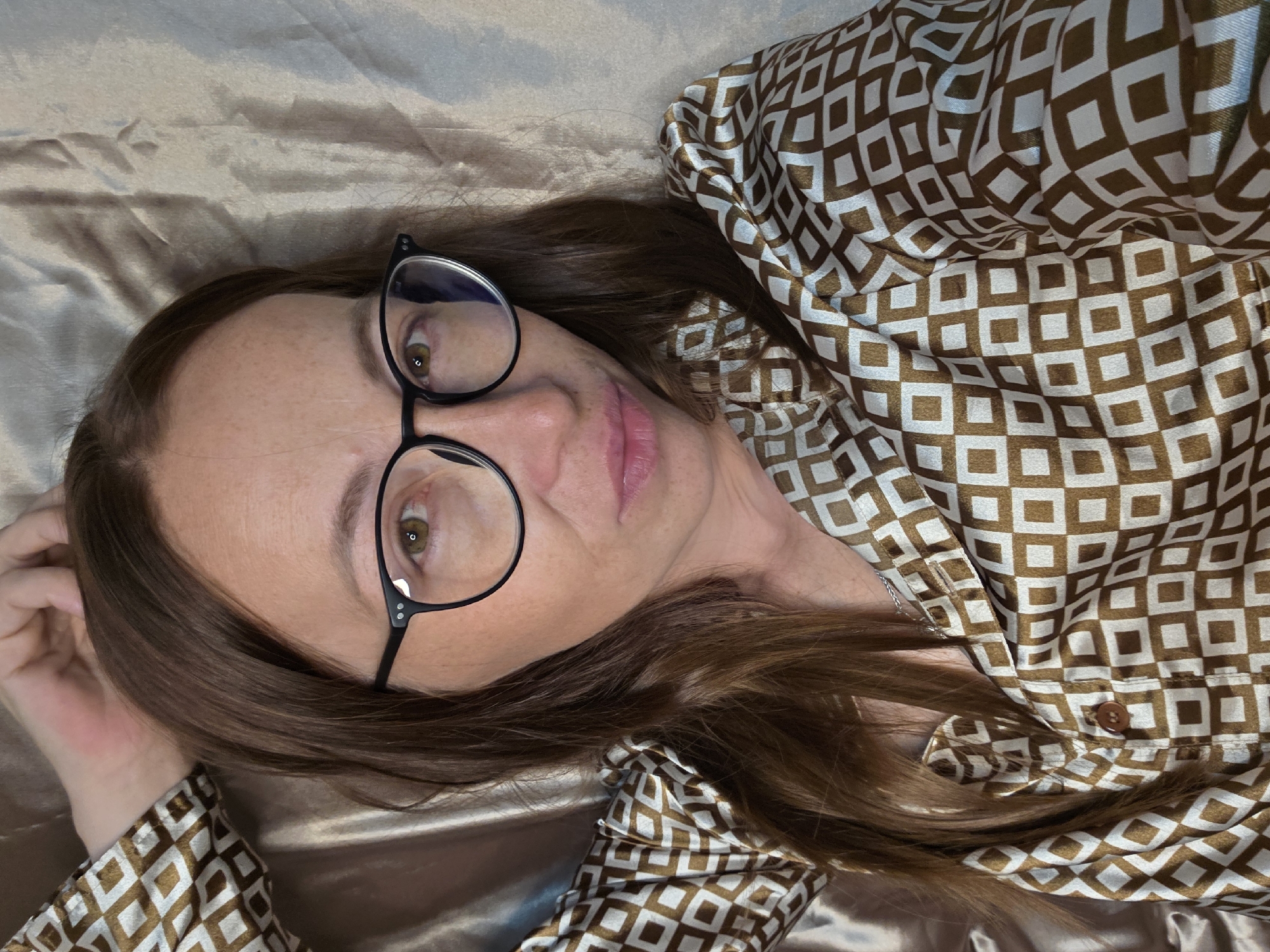 A woman with long, brown hair and glasses is lying on her back on a satin-like surface. She is wearing a brown and white patterned pajama set. Her left arm is raised and resting behind her head. The background is a rumpled, light-colored fabric. The scene appears to be an indoor setting, possibly a bedroom.