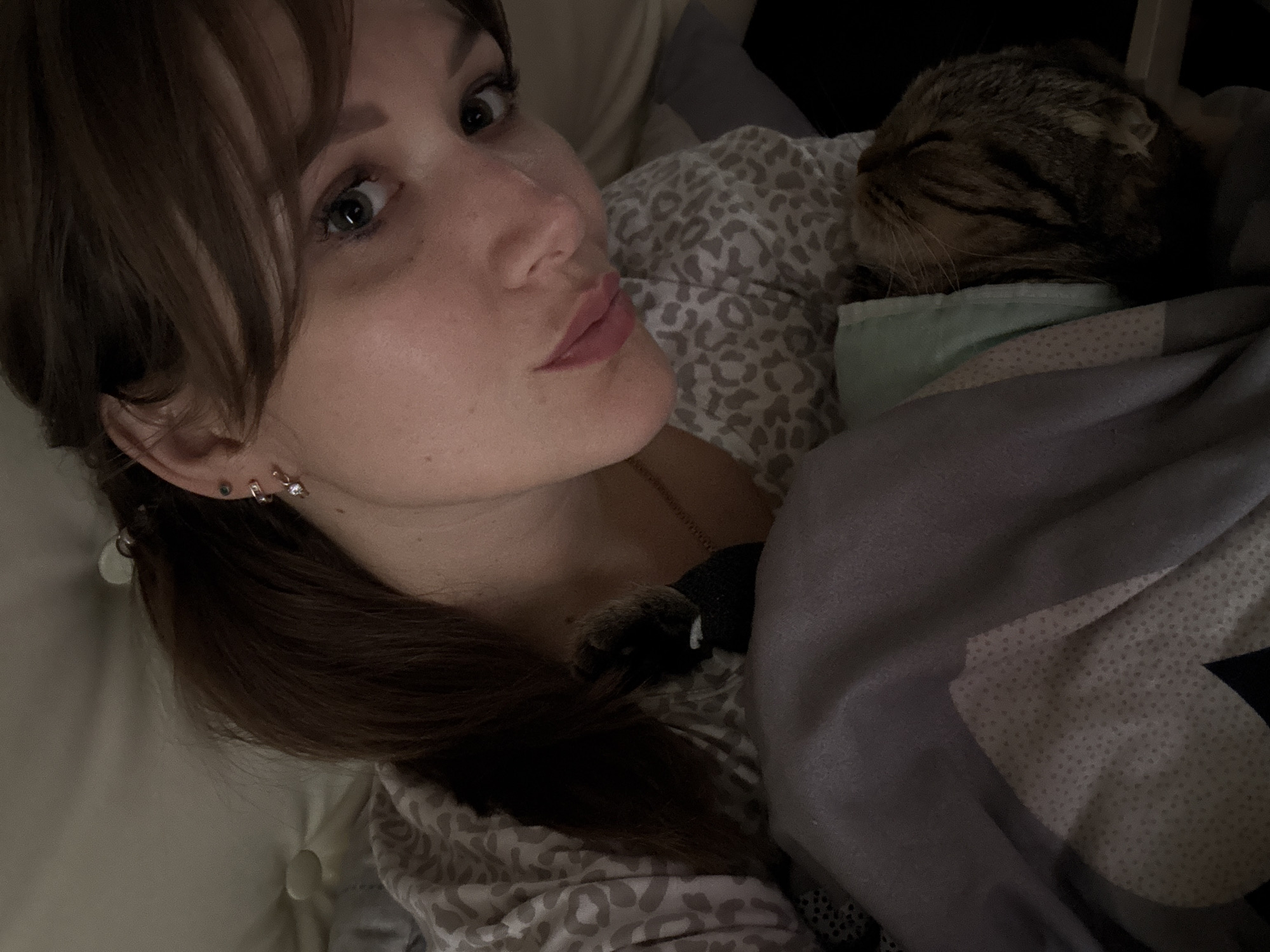 A close-up selfie shows a young woman with brown hair, bangs, and green eyes, wearing a patterned shirt and earrings. She is lying down, with a cat nestled in the blankets beside her. The background appears to be a bedroom, with soft lighting and a hint of a headboard. The image was taken indoors, likely at night, as indicated by the low light conditions. No specific location in Chaikovsky, Russia is identifiable.
