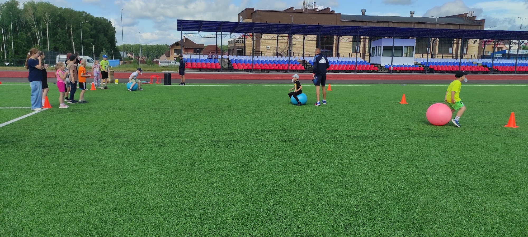 On a bright, sunny day, a group of children and adults are participating in an outdoor event at a sports complex. The scene is set on a vibrant green, artificial turf field, typical of a modern sports facility. In the background, a stadium with tiered seating, colored in red, white, and blue, is visible. A building with a dark, textured facade stands behind the stadium, suggesting an urban or suburban setting. The sky is partly cloudy, adding a dynamic element to the overhead view.
Several children are actively engaged in activities. In the foreground, a child in a bright yellow t-shirt and green shorts is running alongside a large, pink exercise ball. Further to the left, another child, wearing a blue cap and a light blue t-shirt, is seated on a large blue exercise ball, appearing to be in motion. Nearby, an adult, dressed in dark athletic wear with the Adidas logo on the back, stands observing the child.
A line of children and adults are positioned further to the left, seemingly waiting for their turn or observing the activities. They are dressed in casual summer attire, including t-shirts, shorts, and jeans. Orange traffic cones are strategically placed on the field, indicating boundaries or a course for the activities. The overall atmosphere suggests a community event, perhaps a sports day or a children's activity program. The presence of a white van in the distance, along with trees and residential buildings, provides context for the location within the city of Noviy, Russia.