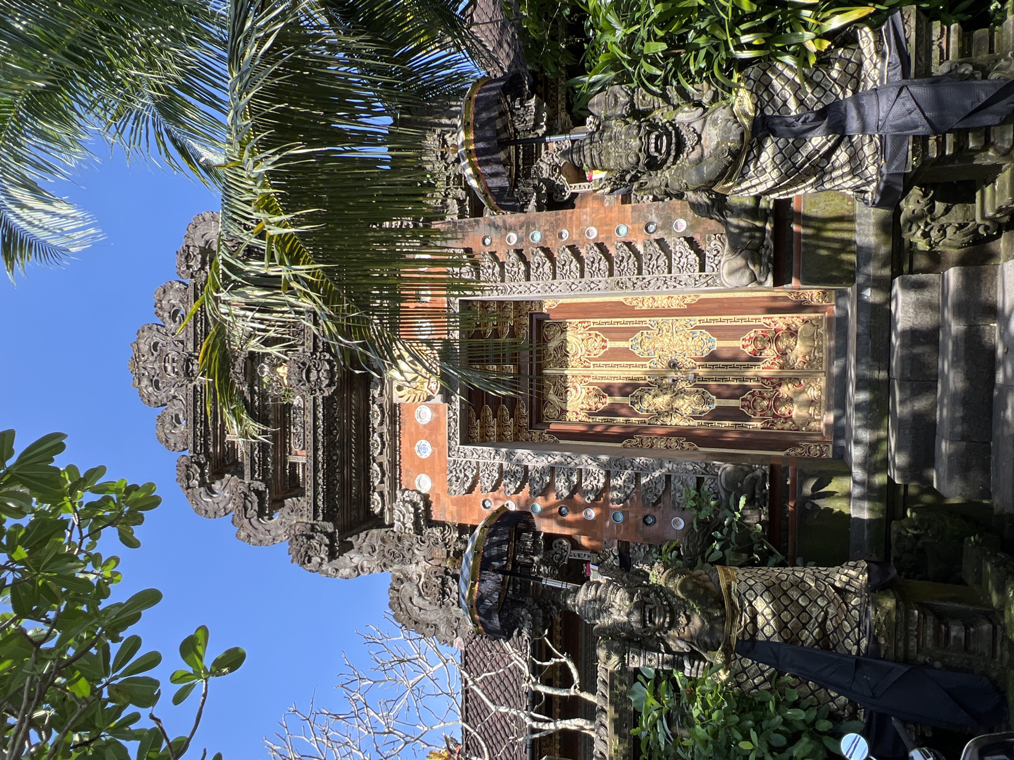 The image shows a highly ornate gate in Petulu, Indonesia. The gate is constructed of carved stone and wood, and is adorned with detailed ornamentation, including rows of decorative discs and intricate patterns. Palm fronds and other greenery partially obscure the gate's upper sections and sides. Two stone statues flank the gate's entrance, draped in patterned fabric. A black umbrella is positioned next to one of the statues. Steps are visible on the right side of the gate. The sky is clear and blue.