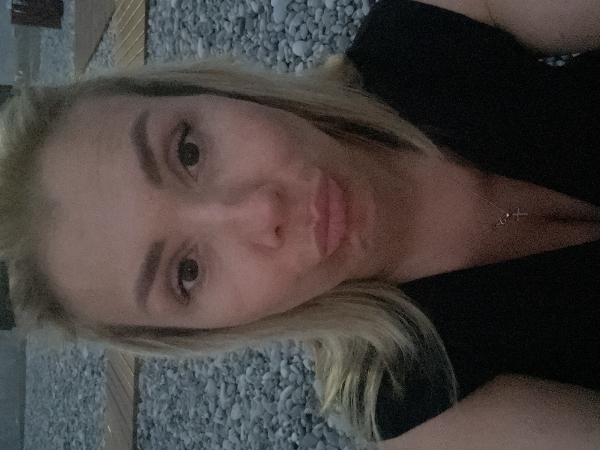 A close-up, selfie-style portrait of a blonde woman with her mouth pursed is captured outdoors. She wears a black dress and a silver necklace with a cross pendant. The background consists of gray pebbles and wooden planks, suggesting a beach or paved outdoor area. The lighting is dim, creating a slightly shadowed effect on her face.