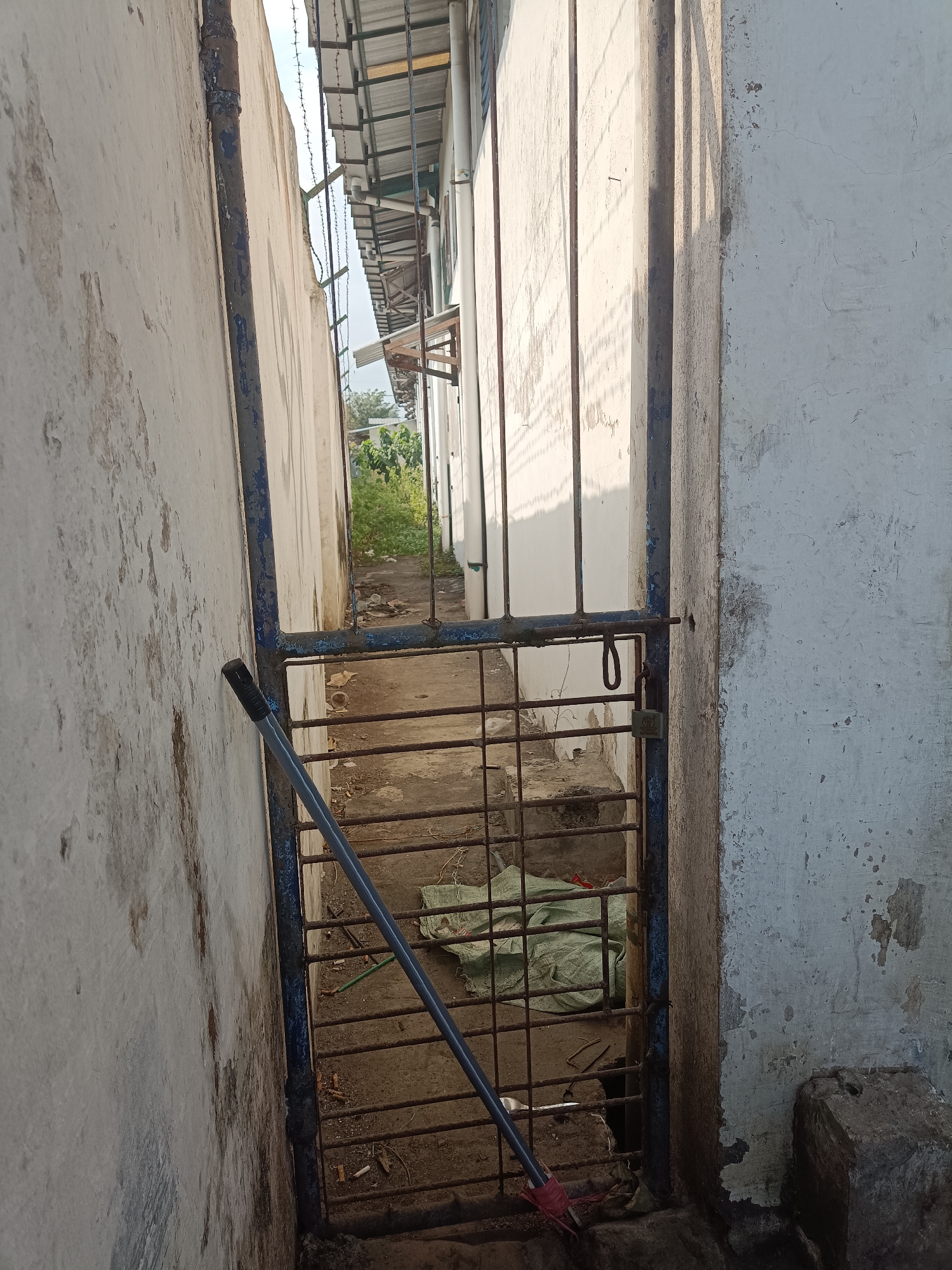 The image shows a narrow alleyway between two buildings, possibly in Jember, Indonesia. The alleyway is partially covered by a corrugated metal roof supported by metal poles. A rusty metal gate with a lock blocks the entrance to a deeper section of the alley. A broom with a blue handle leans against the gate. The walls of the buildings are made of concrete and appear weathered, with peeling paint. The ground is paved with concrete and scattered with debris, including cigarette butts and a green tarp. Sunlight casts shadows across the scene, indicating it is daytime. The overall impression is one of neglect and disrepair.