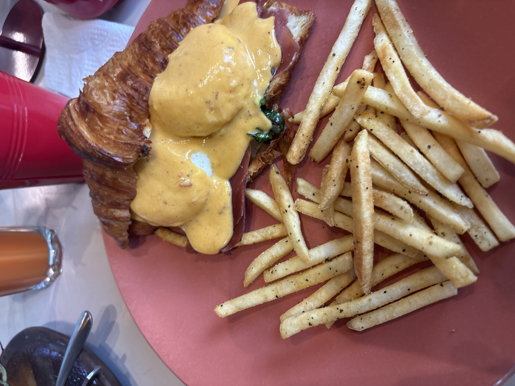 A close-up shot of a plate of food. The main course is a croissant sandwich topped with an egg and a yellow sauce, served with a side of french fries. A glass of juice, a red container and some silverware are on the left. The plate is on a table in a restaurant. The location is Puebla, Mexico.