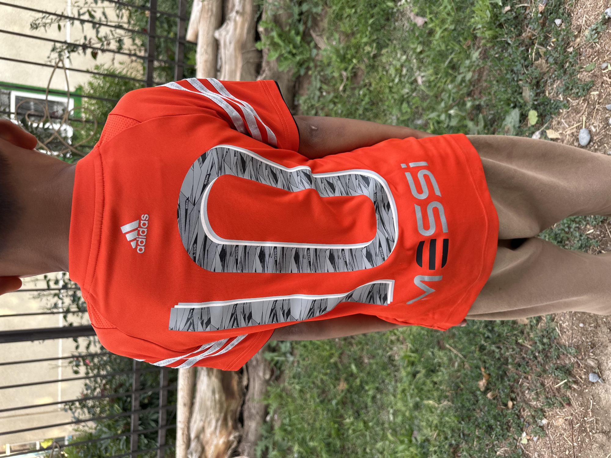 The media file features a young person wearing an orange Adidas t-shirt with a large "10" printed on the back, along with the name "MESSI." The shirt has the signature Adidas stripes on the sleeves. The subject is standing outdoors, as indicated by the presence of grass, dirt, and what appears to be a tree trunk in the background. There is also a metal fence visible on the left side of the image. The weather appears to be overcast or diffused daylight, as there are no strong shadows. The setting suggests a casual outdoor environment, potentially a backyard or a park area. The person's arm is visible, and they are wearing brown pants. The overall tone is casual and sporty. The visible text includes "adidas," "MESSI," and the number "10."