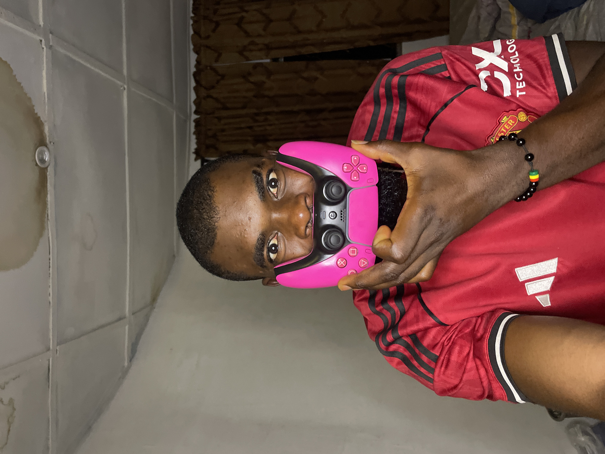 A dark-skinned man, identified as being in Jos, Nigeria, is shown from the chest up, looking directly at the camera. He has short, dark hair and a beard. He is wearing a red Manchester United jersey with black stripes on the sleeves and a white Adidas logo on the left side of the chest. A pink video game controller is held in front of his face, partially obscuring his mouth and nose. He is wearing a black beaded bracelet with red, yellow, and green beads on his left wrist. The background shows a textured ceiling with water stains and a light fixture, as well as brown curtains. The overall lighting suggests an indoor setting, likely at night.