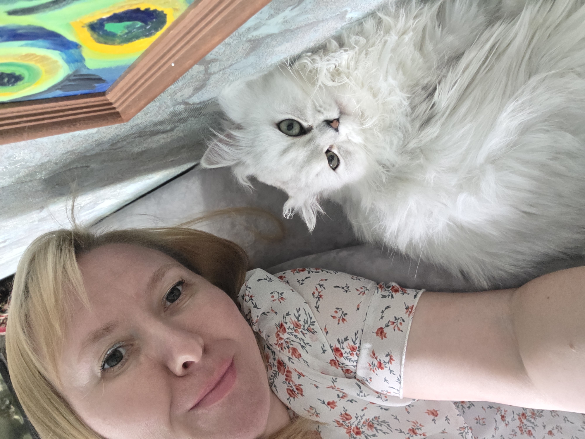 A blonde woman in a floral print shirt lies on a surface next to a fluffy white cat. A framed painting with vibrant blue, yellow, and green colors hangs on the wall behind them. The woman is looking at the camera and smiling, while the cat is looking away from the camera. The scene appears to be indoors.