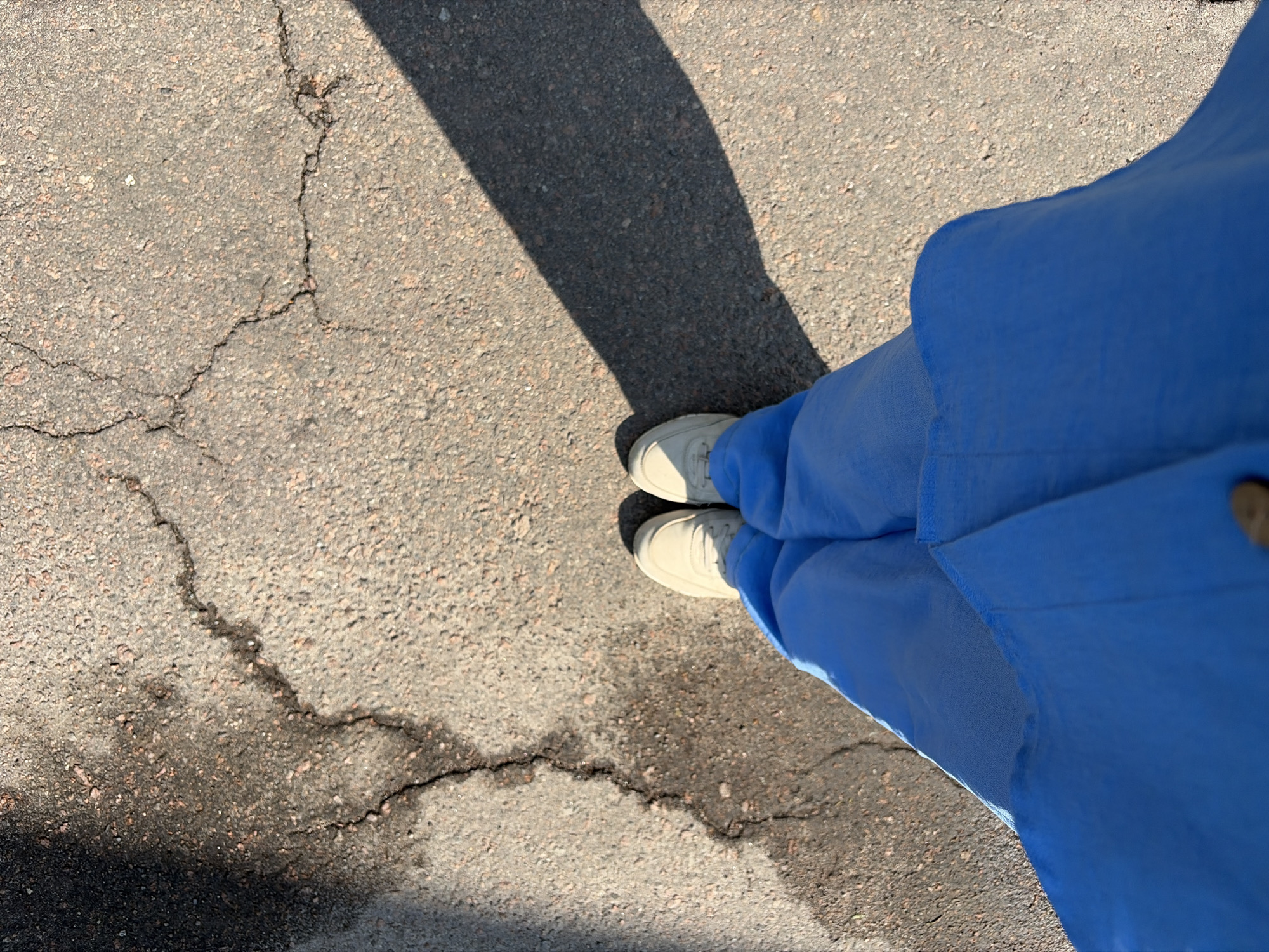 A high-angle, close-up shot captures a person's lower body and feet standing on a cracked asphalt surface. The person is wearing bright blue pants and light-colored sneakers. A sharp shadow extends diagonally across the asphalt from the right side of the frame. The asphalt is grey with visible cracks and some dark, wet-looking patches. The lighting suggests a sunny day.