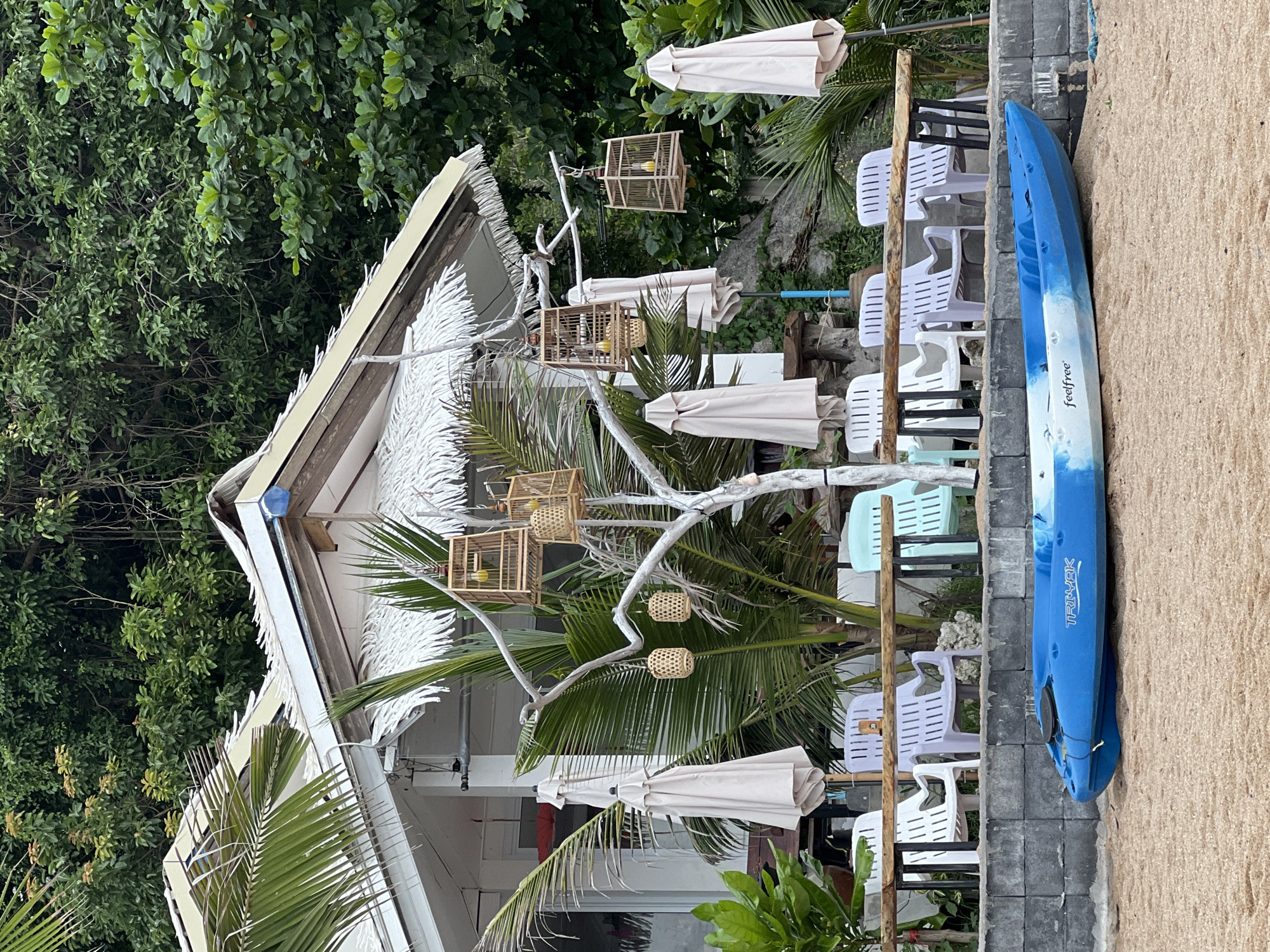 A blue and white kayak rests on the sandy beach in front of a beachside establishment in Baan Chaweng Noi, Thailand. The establishment features a white building with a thatched roof and a white tree sculpture adorned with decorative bird cages and woven hanging lanterns. Several closed, light-colored umbrellas are positioned behind a low retaining wall, which is lined with an assortment of white, purple, and light blue plastic chairs. Lush green foliage, including palm trees, forms a dense backdrop behind the building.