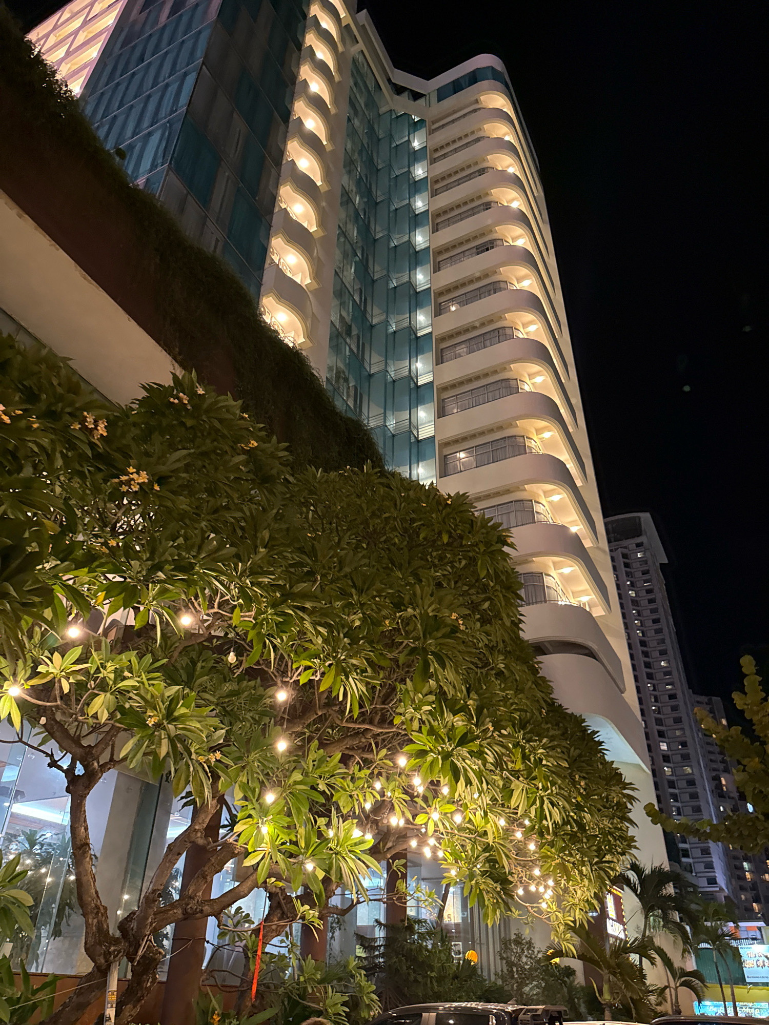 In the city of Nha Trang, Vietnam, at night, a tall, modern hotel building with curved balconies illuminated by warm interior lights dominates the scene. The facade of the hotel on the left is a mosaic of blue-tinted glass panels, creating a striking contrast with the white, rounded balconies on the right. In the foreground, lush green trees with numerous branches are adorned with strings of small, bright lights, casting a festive glow. Some of these trees have small, light-colored flowers. Behind the main hotel, another taller, grey building with rows of lit windows is visible. The night sky is dark, providing a stark backdrop for the illuminated structures and trees. The overall impression is one of a vibrant urban night scene, possibly a hotel entrance or outdoor seating area, designed for evening ambiance.