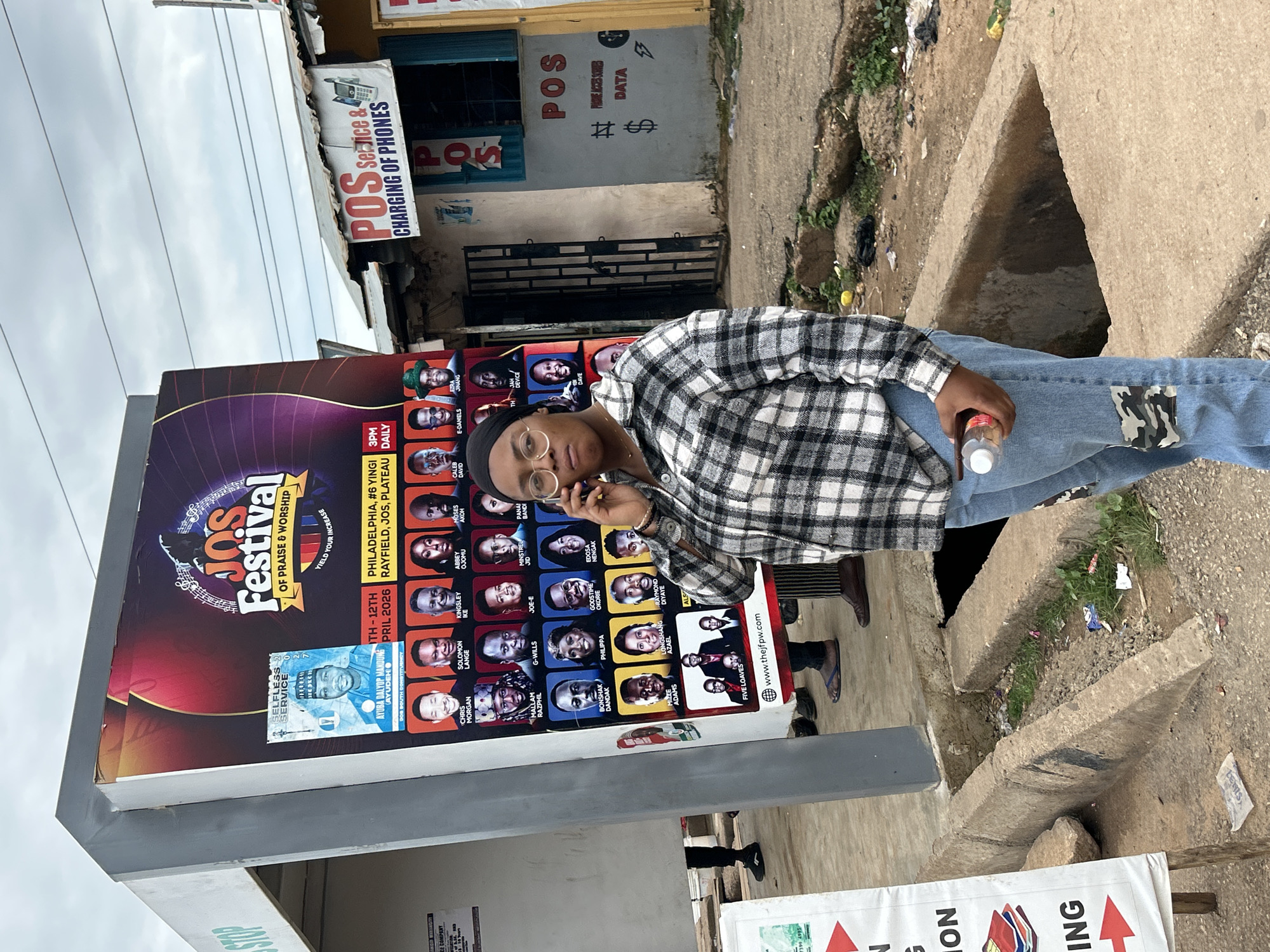 A woman with a black headscarf and round glasses stands on a street in Jos, Nigeria, talking on her cell phone. She is wearing a black and white plaid shirt and light blue jeans with camouflage patches on the knees. She holds a clear plastic bottle with a red cap in her right hand.
Behind her, a large billboard advertises the "JOS FESTIVAL OF PRAISE & WORSHIP" with the tagline "YIELD YOUR INCREASE." The billboard features portraits of various individuals and lists event details: "6TH - 12TH APRIL 2026, PHILADELPHIA, #6 YINGI RAYFIELD, JOS, PLATEAU." To the right of the billboard is a small shop with a sign that reads "POS Service & CHARGING OF PHONES."
The environment appears to be a somewhat unkempt urban setting. There is a drainage ditch in the foreground filled with debris and overgrown vegetation. The ground is paved with concrete and dirt. The sky is overcast. Several other individuals are visible in the background, some standing near the billboard and others near the shop.