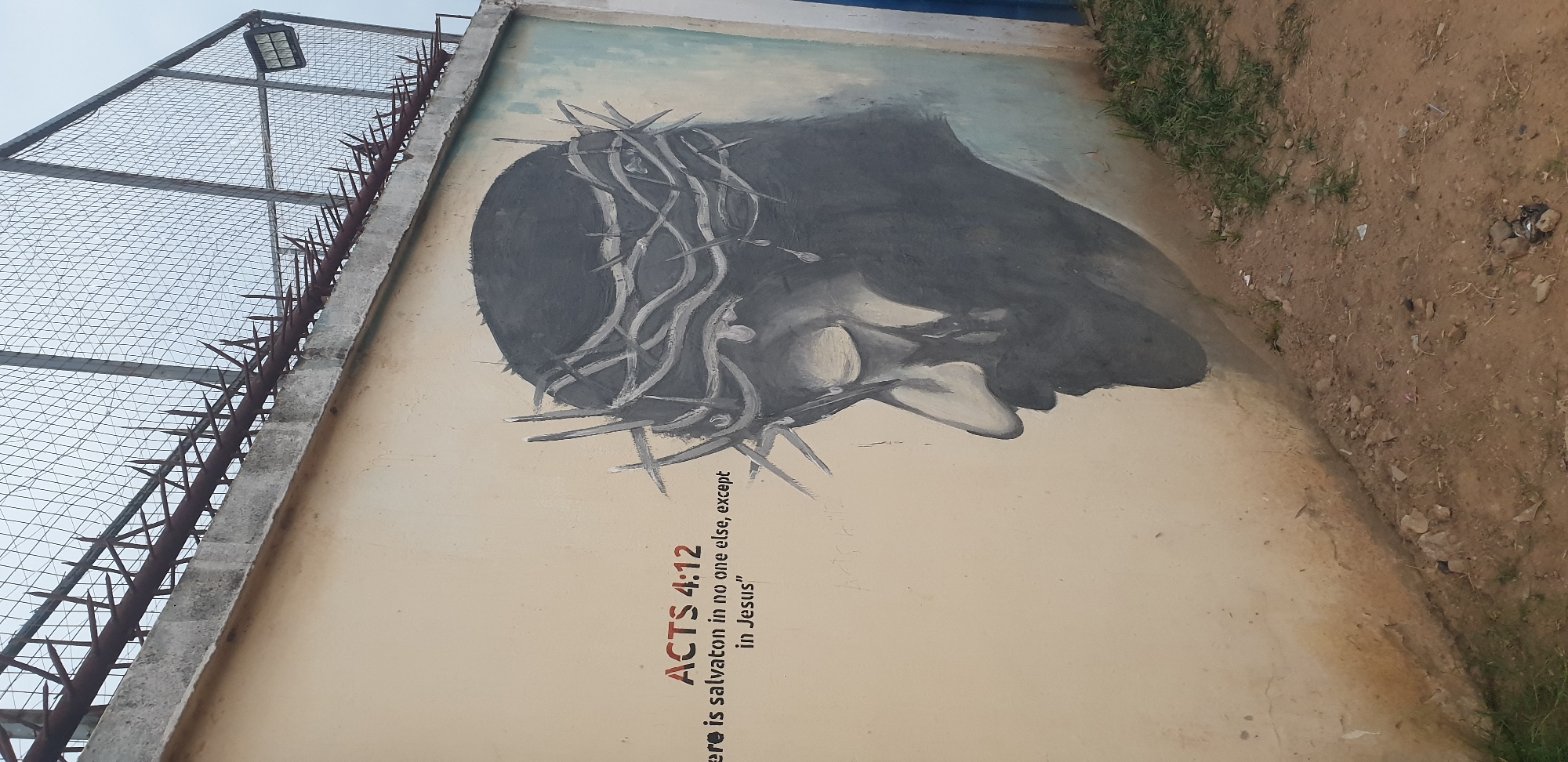 The image captures a mural on a concrete wall in Sabon Tasha, Nigeria. The mural depicts a stylized portrait of Jesus Christ wearing a crown of thorns, rendered in shades of gray and black. To the left of the mural, the text "ACTS 4:12" is prominently displayed in red, followed by the quote "There is salvation in no one else, except in Jesus" in black. The wall itself is painted a light beige color, with some visible scuff marks and imperfections. Above the wall, a chain-link fence with barbed wire is visible, suggesting the location might be a secure or fortified area. The ground in front of the wall is a dirt path with scattered small stones and a few patches of green grass. The sky appears overcast.