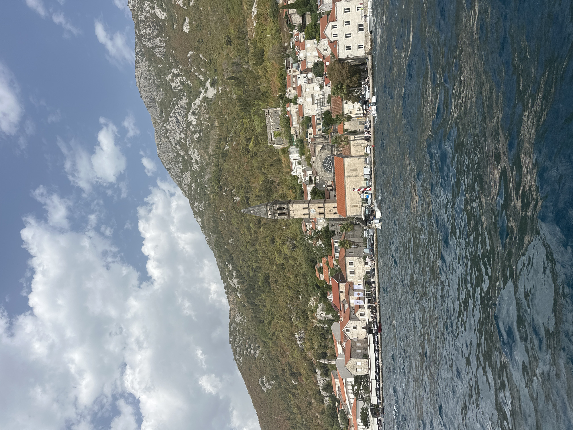 A medium shot captures a view of Perast, Montenegro, from the water. The town is nestled against a steep, rocky mountainside covered in scattered green foliage. A dominant bell tower with a clock face rises from the center of the town, indicating it is likely part of a church complex. Buildings with red-tiled roofs and stone facades line the waterfront, with some appearing to be restaurants or shops. Several boats are visible in the water and docked along the shore. The water is a deep blue with visible ripples and waves, suggesting a slight breeze. The sky is a mix of blue with fluffy white clouds. The scene is bathed in natural daylight.