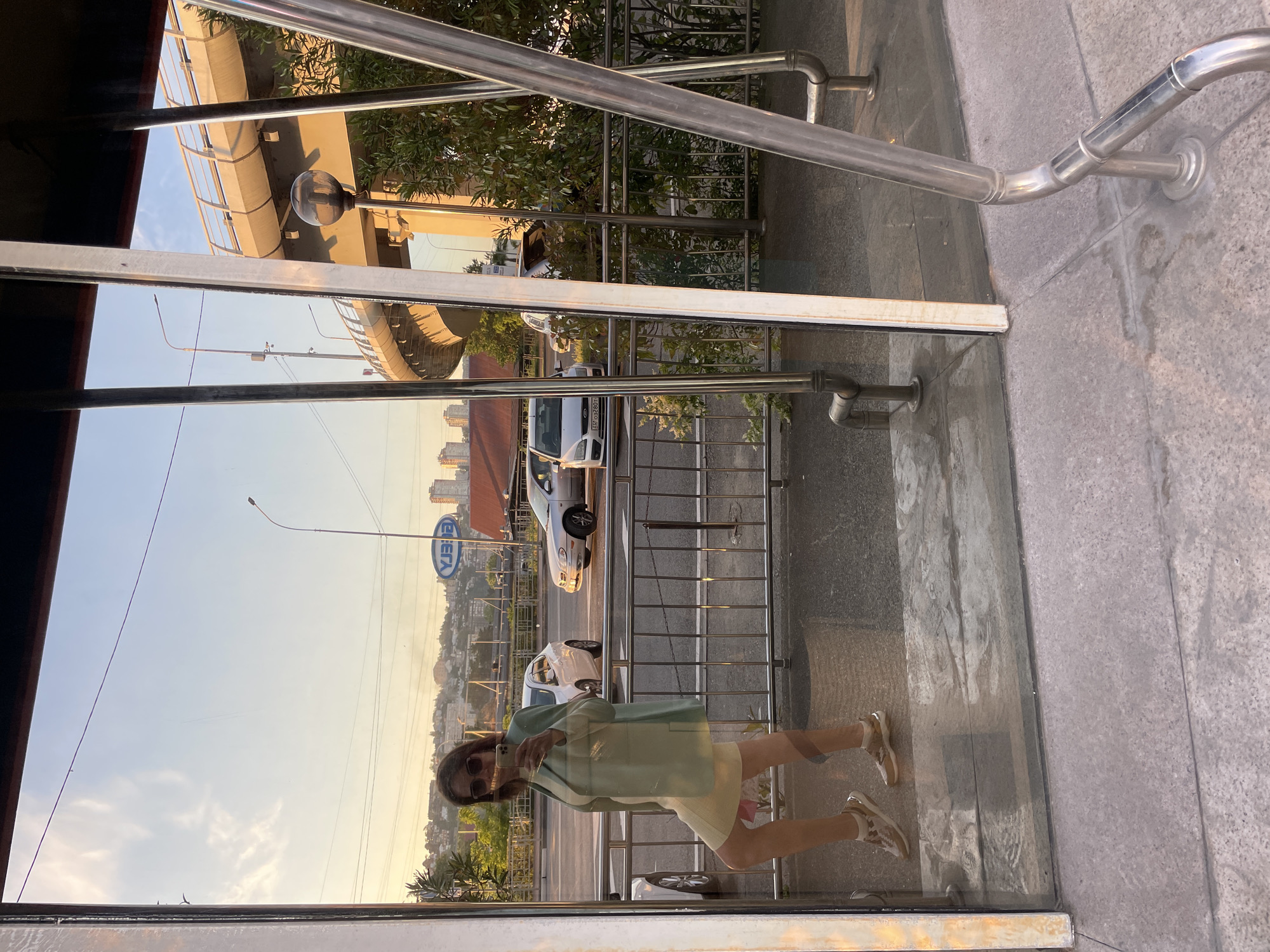 A vertically oriented image captures a person taking a mirror selfie through a glass panel, revealing an urban outdoor scene in reflection. A young woman, dressed in dark sunglasses, a light green jacket, a light-colored skirt or dress, and light-colored athletic shoes, holds a smartphone to capture her reflection.
The background, seen through the glass and as a reflection, depicts an urban environment during what appears to be the golden hour of late afternoon or early evening, characterized by warm, soft lighting. A street or road with vehicular traffic is visible, including a white SUV/sedan and a silver car. Prominent metallic railings run along the glass barrier and also form parts of an elevated road or bridge structure. Green trees and foliage are interspersed throughout the scene.
In the distance, a city skyline stretches across a landscape with rolling hills or mountains under a hazy sky. A street light fixture is also visible. On a blue sign, reflected and therefore appearing reversed, the text "CIETA" is discernible. The setting suggests a modern urban area, possibly an observation deck or a pedestrian overpass in a city like Sochi, Russia.