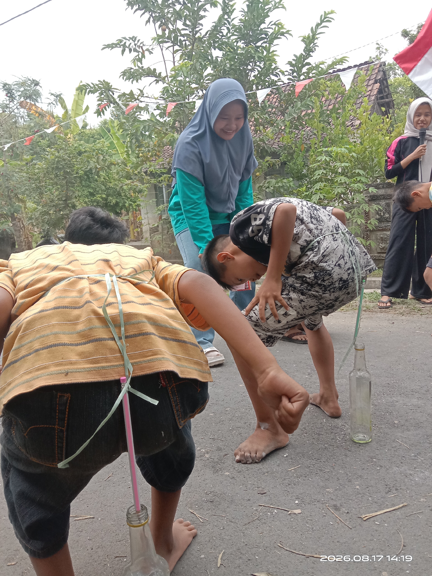 The image shows children participating in a game in Banyuarang, Indonesia. Two young boys are bending over, each with a straw attached to their clothing and extending into a glass bottle on the ground. They appear to be trying to blow air through the straw to move the bottle. A young girl in a blue hijab is standing behind one of the boys, smiling and watching the game. Another girl in a white hijab is standing to the right, holding a microphone. In the background, there are trees, houses, and festive red and white flags strung across the area. The ground is paved with asphalt. A timestamp on the image reads "2026.08.17 14:19".