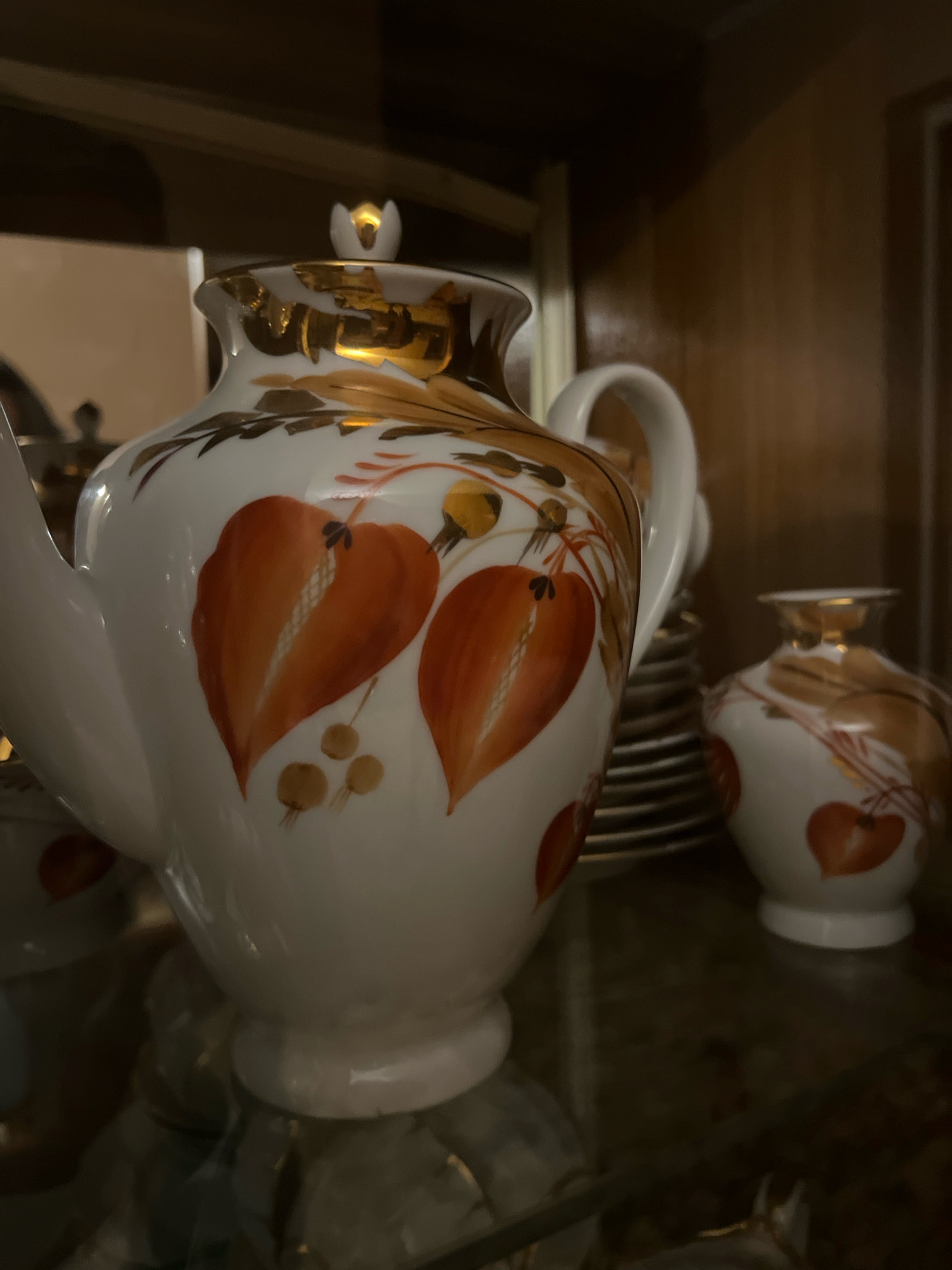 A close-up, eye-level shot captures a white ceramic teapot with an ornate gold and orange leaf and fruit design. The teapot is adorned with two large, stylized orange leaves with prominent veins, and smaller clusters of orange berries. A gold decorative band circles the rim of the teapot and accents the lid. To the right of the teapot, a smaller, matching ceramic vase with a similar gold and orange floral pattern is visible. Both items are displayed on a reflective surface, possibly a shelf, with a dark wooden background. The lighting is dim and focused, creating a warm and intimate atmosphere. The scene appears to be a still life arrangement of decorative tableware.