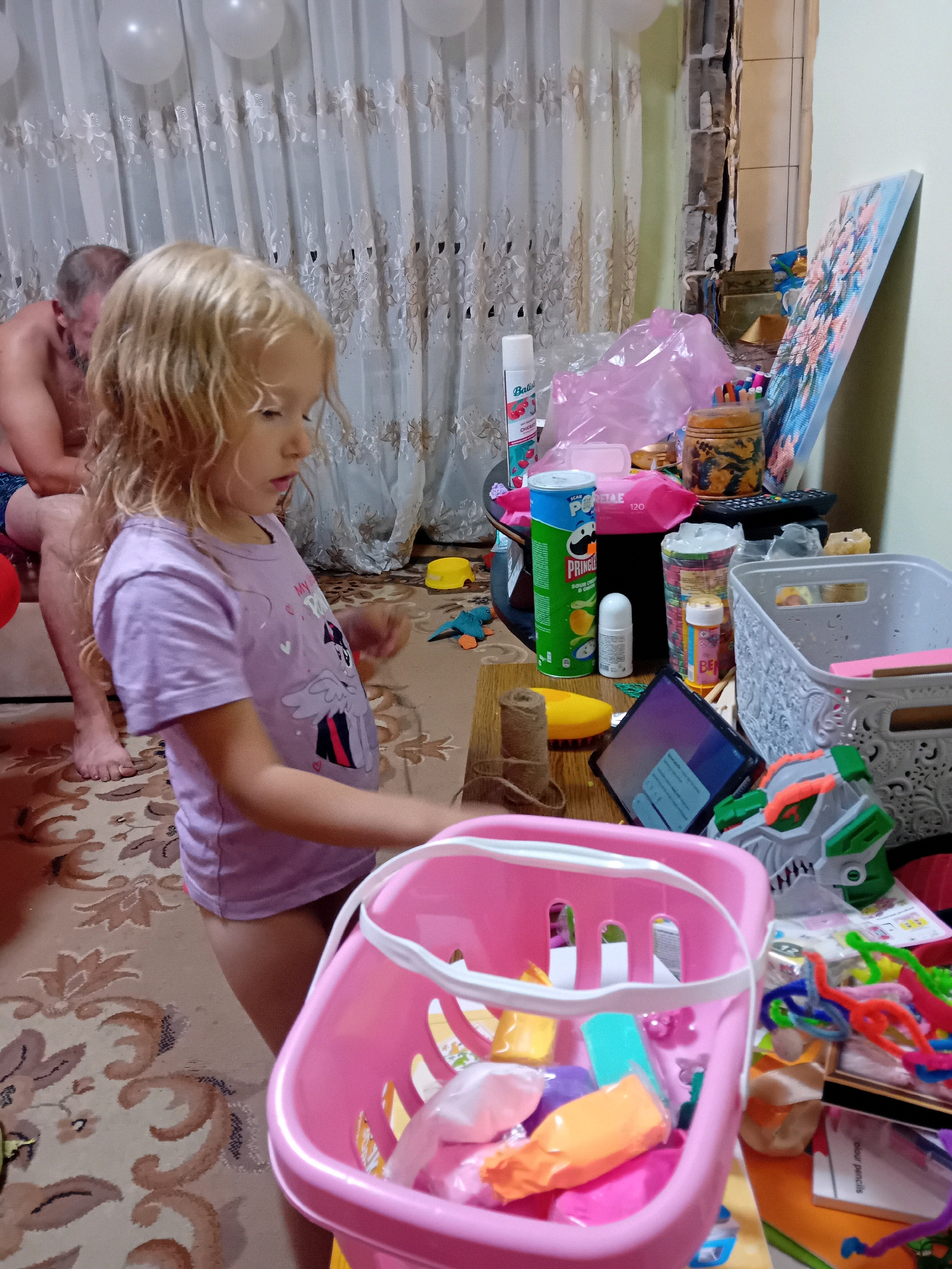 A young girl with blonde, curly hair is standing in a room with a man in the background. The girl is wearing a light purple t-shirt and is reaching into a pink plastic basket filled with various colorful objects, including what appears to be modeling clay. The room appears to be a domestic setting, possibly a living room or play area, with a patterned carpet, curtains, and a table cluttered with items like Pringles, a tablet, and craft supplies. There are balloons visible in the background, suggesting a celebration. The overall impression is one of everyday life with a child engaged in play.