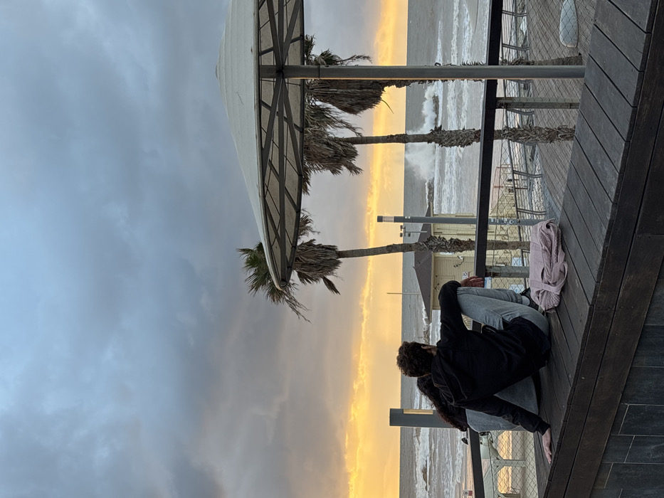 The image depicts a serene coastal scene in Tel-Aviv, Israel, likely at sunset or late afternoon. A dramatic sky features a vivid golden-orange band along the horizon where it meets the sea, contrasting sharply with the dark, cloudy expanse above.
In the foreground, two people are seated on a wooden deck or promenade, facing away from the camera and towards the sea. One individual, with dark curly hair, is predominantly visible, wearing dark clothing and light-colored trousers, sitting with their legs drawn up. The other person is partially obscured beside them. A light pink blanket or clothing item is folded neatly on the wooden deck next to them.
The setting includes a prominent large white, conical shade structure, possibly a gazebo or umbrella, supported by metal framework. Several palm trees with somewhat sparse fronds are visible, extending upwards near this structure. Beyond the wooden deck and its railings, the sea appears turbulent with white-capped waves crashing, indicating windy conditions. A small building or structure can be seen faintly in the distance along the coastline. The overall mood is contemplative, with the individuals seemingly observing the striking natural phenomenon. There is no visible text in the image.