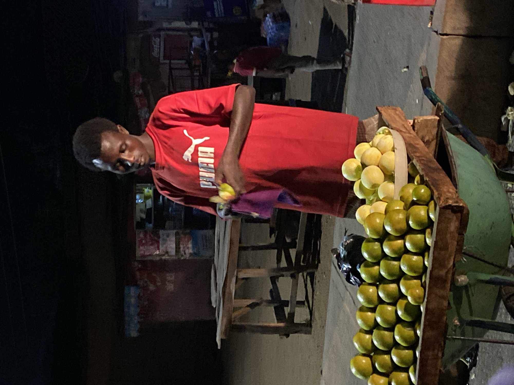 A young man is depicted in a street or market setting in Bukuru, Nigeria, likely at night. He stands focused, looking down at his hands, where he appears to be holding a piece of fruit and possibly wiping it with a purple cloth or rag. He is wearing a red short-sleeved t-shirt with the white "PUMA" logo prominently displayed on the front.
To his right, a rustic wooden cart or stand holds a large display of neatly stacked fruit, predominantly green and yellow-green, resembling oranges or limes. The fruit is arranged in rows within compartments of the wooden structure, with some lighter yellow fruit visible at the top. The cart has some green painted sections.
The background is dark and blurred, suggesting artificial lighting is the primary light source, illuminating the man and his fruit stand. Faint outlines of buildings or market stalls can be seen behind him, including what might be a red "Coca-Cola" sign in the upper background of the main image and some blurry shelves or structures. The overall scene conveys the atmosphere of an evening street vendor or market stall. The man's expression is neutral and concentrated on his task.