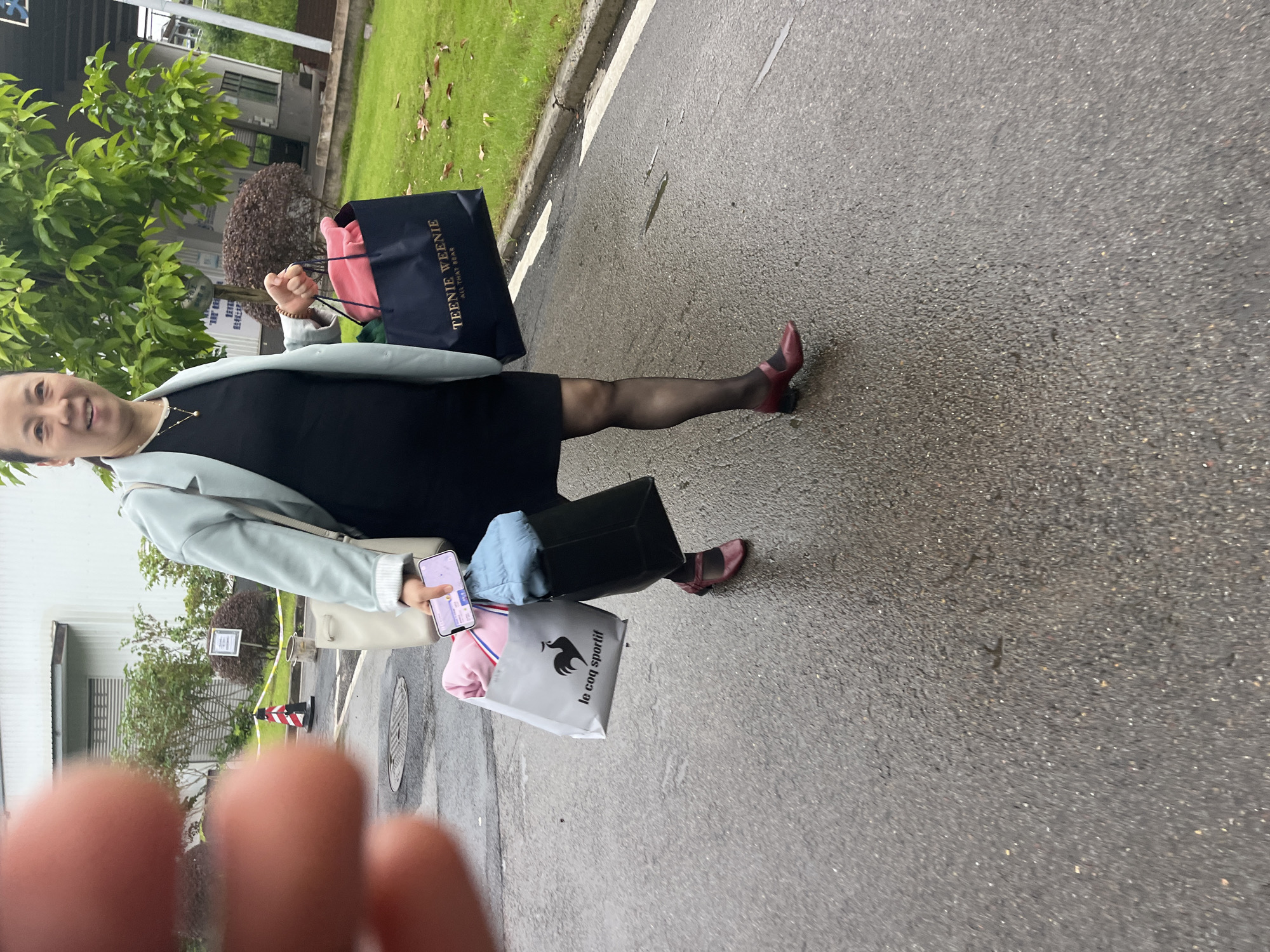 A woman is walking on a wet asphalt road in front of a building that appears to be a factory or warehouse. She is carrying several shopping bags. The woman is wearing a light blue blazer over a black dress, black pantyhose, and dark red mary jane shoes. She is holding a smartphone in her right hand, with the screen facing outwards. The shopping bags include a dark navy bag with "TEENIE WEENIE ALL THAT BEAR" printed on it, a light gray bag with a rooster logo and "le coq sportif" printed on it, and a white tote bag. Pink and light blue clothing items are visible inside the shopping bags. The background shows greenery and a building with windows. A striped traffic cone and yellow caution tape are visible to the left of the woman. The presence of the building and the style of the traffic cone suggest an industrial or commercial area. It is possible this scene is located in the city of Jiangjin, China, given the context provided.