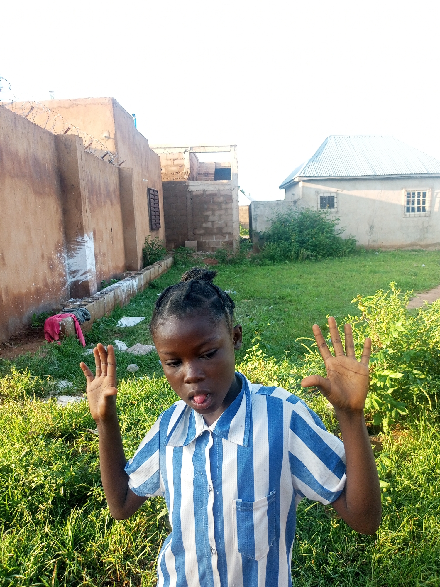 A young girl with braided hair, wearing a blue and white striped shirt, poses with her hands up and tongue sticking out in a grassy yard. Behind her, a textured brown wall with barbed wire runs along the perimeter of a property. Adjacent to the wall is a long concrete trough, and further back, two buildings are visible: one appears to be constructed with bricks and cement, while another has a light-colored metal roof. The overall setting suggests a residential area in Tempure, Nigeria, possibly a schoolyard or a community space. The lighting indicates it is daytime, with sunlight casting shadows on the buildings.