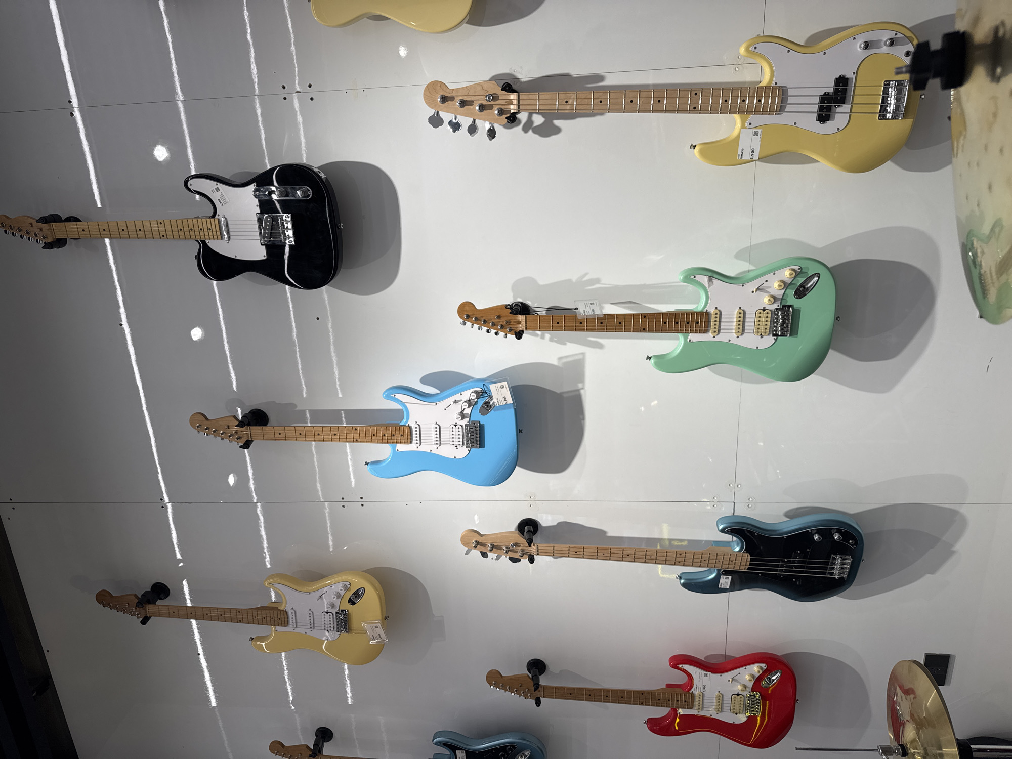 This media file captures a vibrant display of multiple electric guitars and basses, meticulously mounted on a clean, white wall, likely within a music store or showroom in Davao City, Philippines. The instruments are arranged vertically in rows, creating a visually striking exhibit.
In the top left, a black Telecaster-style electric guitar is prominently displayed. Below it, a light yellow Stratocaster-style electric guitar is visible. Further down, a striking red Stratocaster-style guitar is shown. On the right side of the frame, from top to bottom: a light yellow Precision Bass (P-bass) style instrument is visible, featuring a white pickguard. A price tag on this instrument clearly reads "P 9,900". Below the yellow bass, a mint green Stratocaster-style guitar with a white pickguard is displayed. Following this, a light blue Stratocaster-style guitar, also with a white pickguard, is mounted. Below that, a dark blue or teal Precision Bass style instrument, with a black pickguard, is shown. Parts of other instruments, including a light yellow electric guitar and a light blue electric guitar, are also visible at the top and bottom edges of the frame, respectively.
The wall is brightly lit by strong, vertical strip lighting, which casts distinct shadows of each instrument, adding depth to the display. Small, almost indistinguishable price or product tags are attached to several guitars. In the bottom right corner, a portion of a golden cymbal and a drum stand are visible, suggesting a broader musical instrument retail environment. There are no people present in the image, focusing entirely on the instruments themselves.