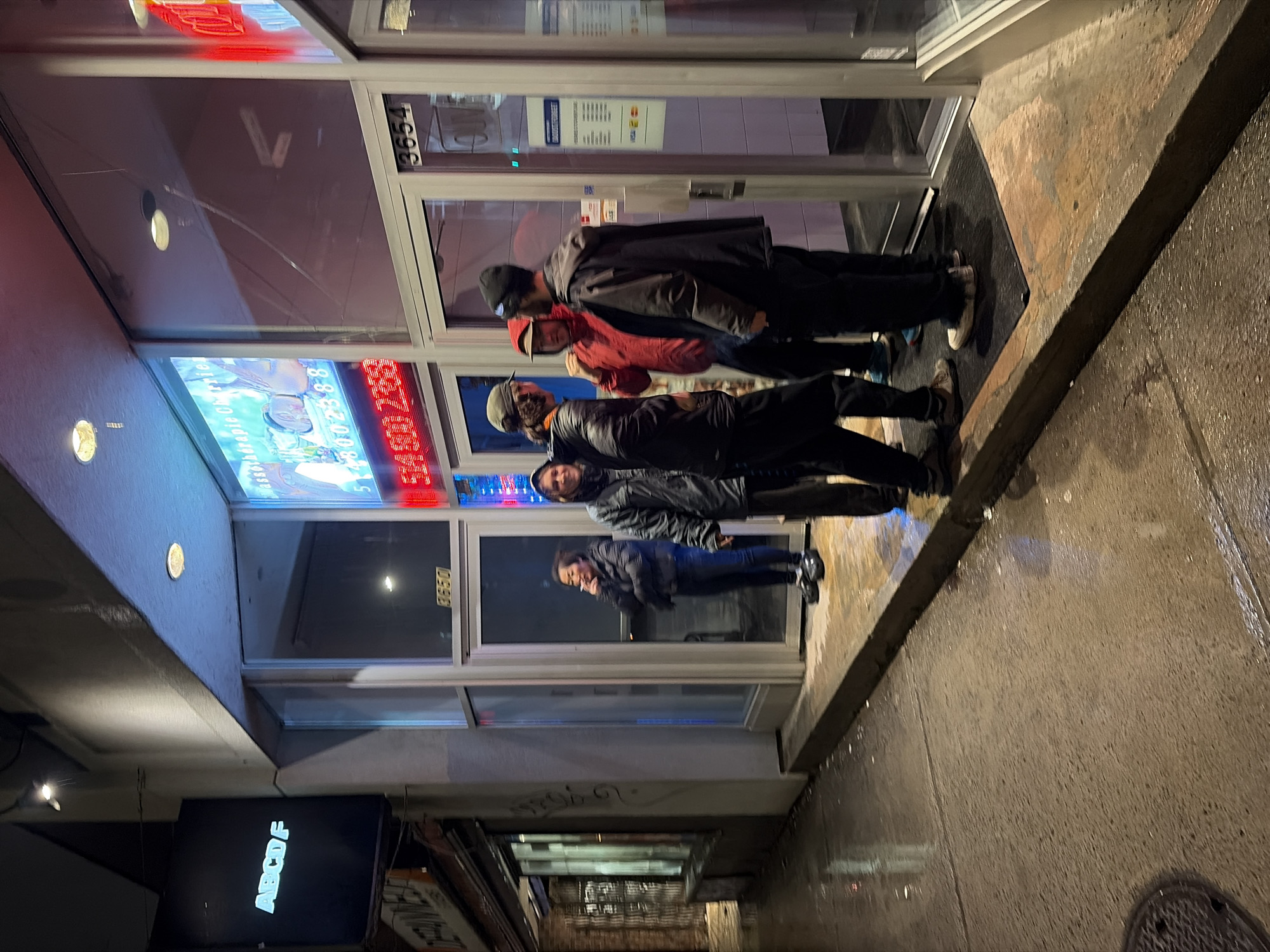 A nighttime street scene shows five people standing in front of a storefront, with one person smoking a cigarette. The storefront is located at 3654 on a street in Montreal, Canada. The group appears to be waiting, with the first three individuals closest to the door. The sidewalk is wet, suggesting recent rain. A business sign with the letters "ABCDE" is visible in the lower-left corner, indicating a commercial establishment nearby. The storefront has a colorful advertisement display and various notices on the windows.