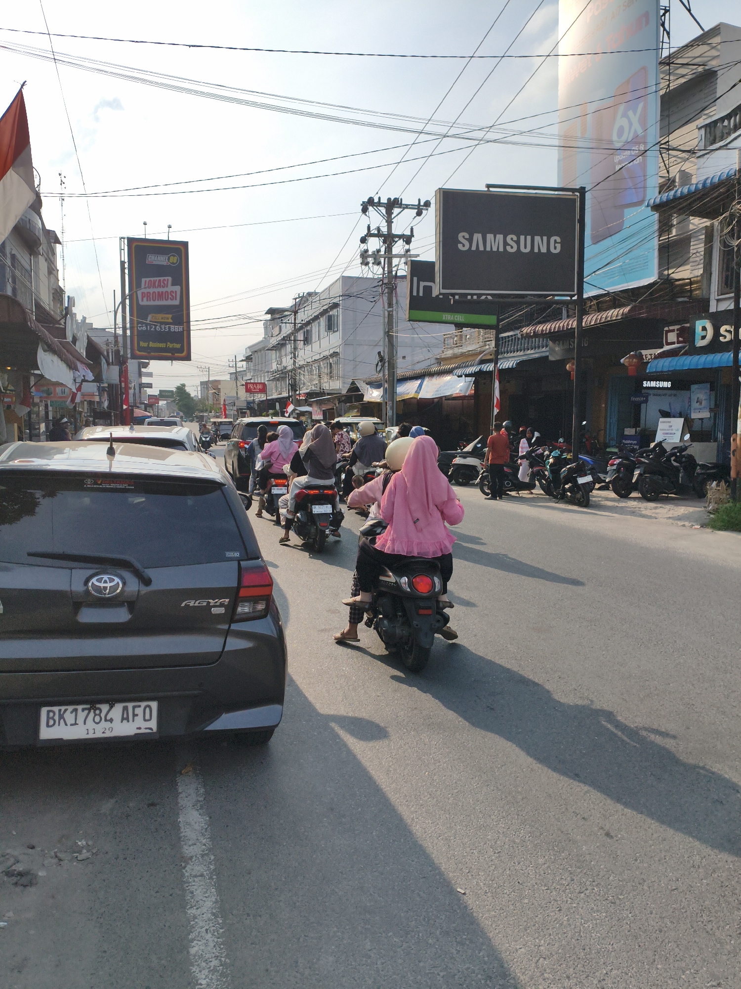 A street scene in the city of Tebing Tinggi, Indonesia, shows moderate traffic during daylight hours. The image is captured from behind a dark gray Toyota Agya, with its license plate reading "BK 1784 AFO 11 29". In front of the car, several motorcycles are in motion, carrying passengers, some of whom are wearing headscarves. A Samsung advertisement is visible on a large billboard above a row of shops. The buildings along the street appear to be shops and commercial establishments. The sky is partly cloudy, and shadows indicate the sun is casting long shadows on the asphalt road. The overall impression is a typical urban environment with daily commutes and commercial activity.
