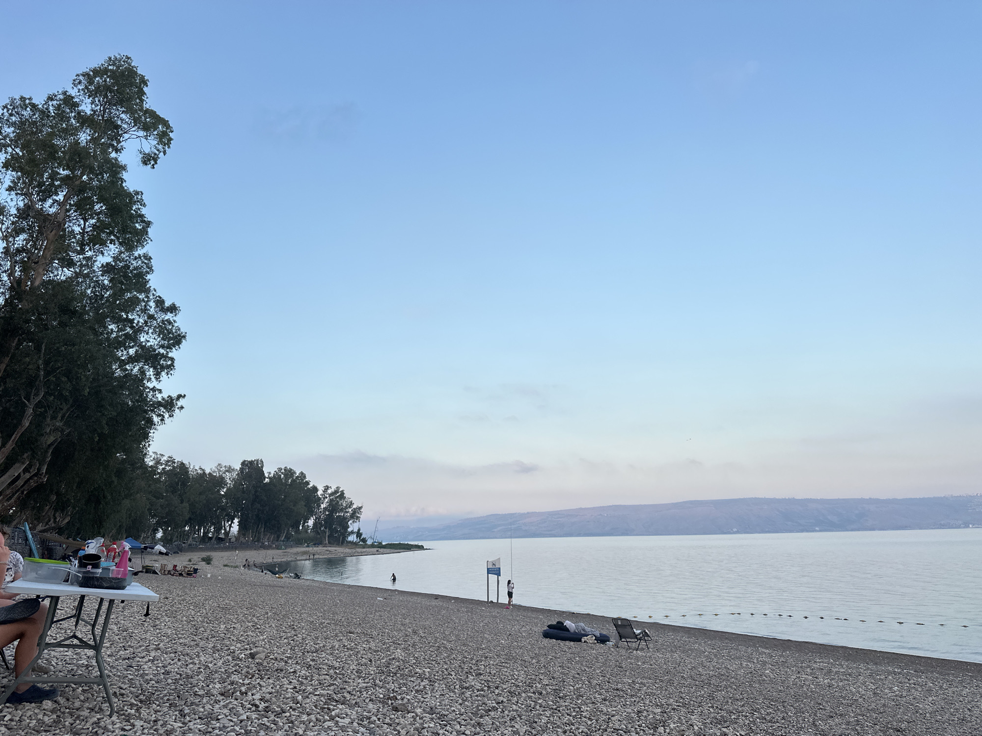 The image depicts a pebble beach on the shore of the Sea of Galilee, located in the Emek HaYarden Regional Council, Israel. The sky is a clear, pale blue with a hint of pink near the horizon, suggesting either dawn or dusk. A calm body of water stretches out, meeting a distant, hazy mountain range.
On the left side of the frame, a large tree with dark green foliage stands prominently, casting shadows on the beach. Beside the tree, a portable table is set up with various items, including bowls and containers, suggesting a picnic or camping setup. A person's legs are visible, seated next to the table.
Further along the beach, several other people are visible, some walking or standing near the water. There are also some tents and beach chairs scattered across the shoreline, indicating a recreational area. A signpost stands near the water, and a line of buoys marks a boundary in the sea. The beach is covered in small, grey and white pebbles. The overall atmosphere is serene and tranquil.