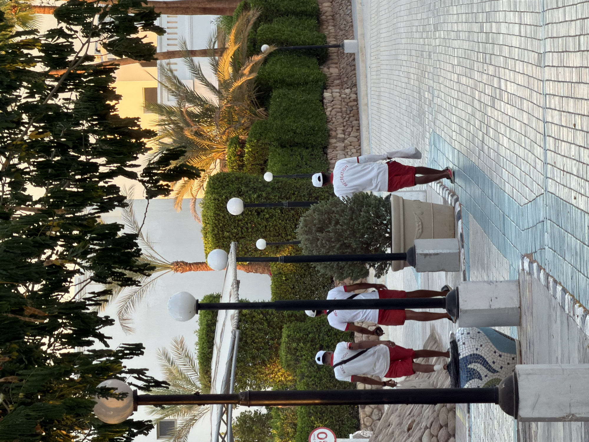 In a scene set in Sharm El Sheikh, Egypt, several individuals dressed in white shirts with red trim and red shorts are walking along a paved walkway. They appear to be staff members, possibly lifeguards or resort employees, as indicated by the word "RECREATION" visible on the back of one of their shirts. The time of day appears to be late afternoon or early evening, with warm golden light illuminating the palm trees and the buildings in the background. The weather seems pleasant, conducive to outdoor activities.
The walkway is bordered by neatly trimmed green bushes and interspersed with white globe lights on black poles. In the background, a building with a light-colored facade and palm trees suggest a resort or hotel environment. The walkway itself is paved with light-colored rectangular tiles, with a decorative blue and white tiled border on one side. The individuals are walking in the same direction, suggesting a patrol or a duty. Their posture is relaxed, and no particular emotions are discernible from this distance. The overall impression is one of a well-maintained, tranquil outdoor space.